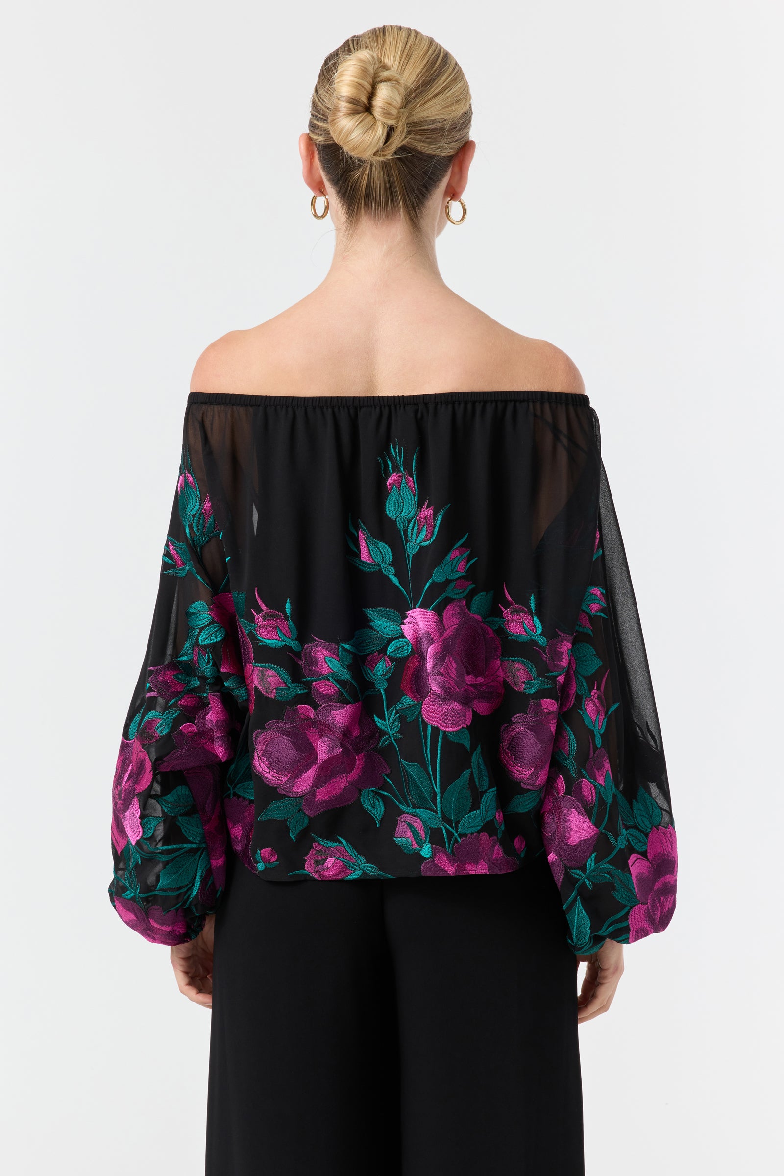 A woman with blonde hair in a bun and gold hoop earrings models the Soho 2 Embroidered Top—an off-the-shoulder black blouse with sheer sleeves and bold pink and green floral embroidery—facing away from the camera.