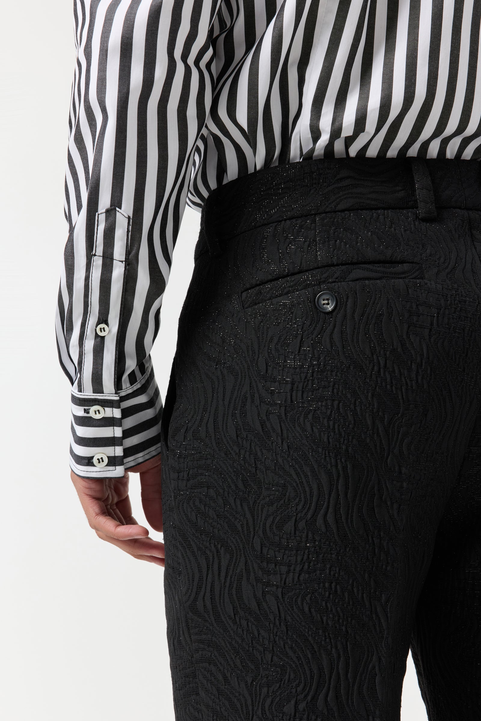 A person is shown from the waist down wearing the Clyde Slim Trouser - Metallic Groove, styled with a black and white vertically striped shirt and their left arm relaxed by their side.