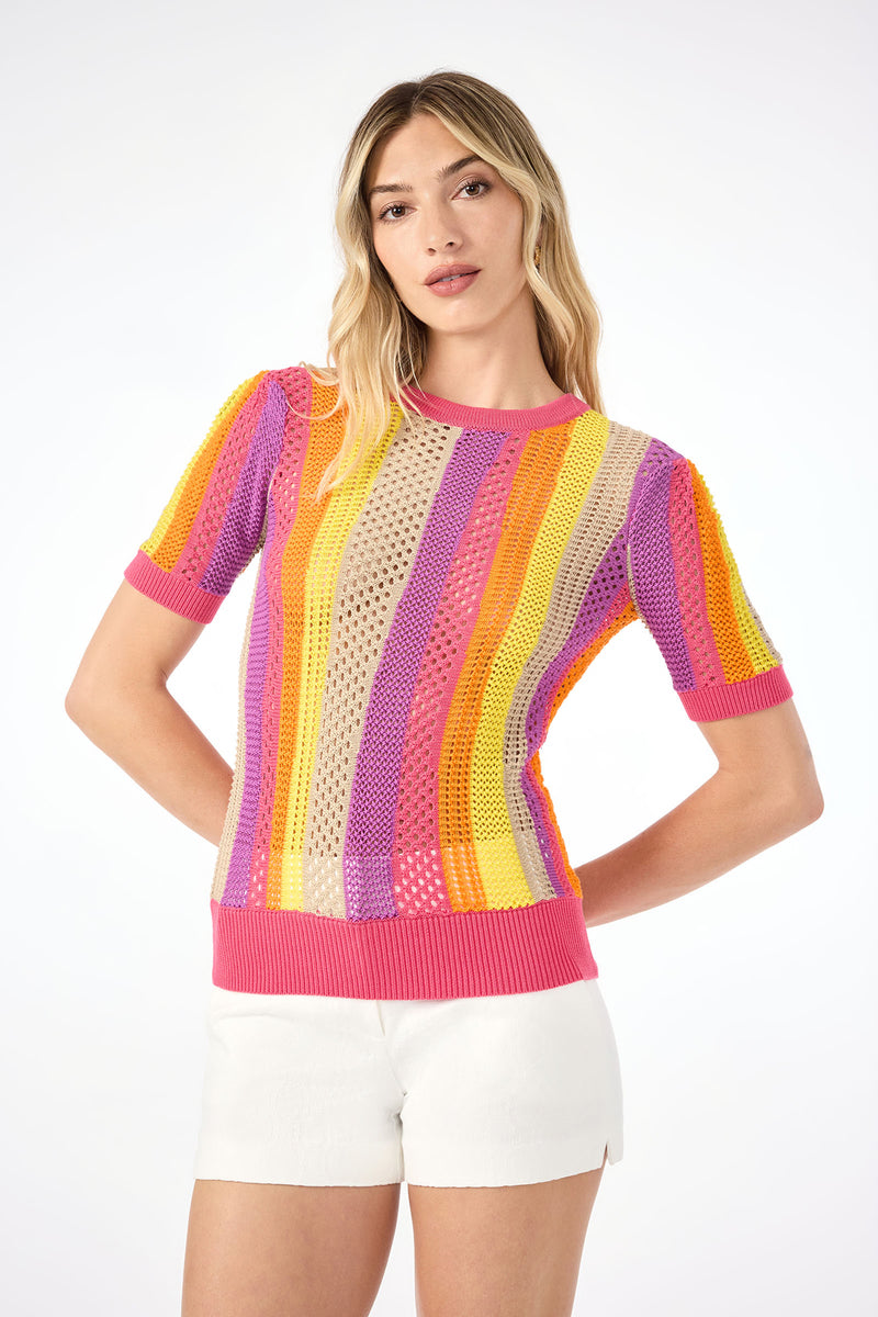 A woman models the Marbella Short-Sleeve Pullover in Eden Roc Rose, featuring vibrant stripes, paired with white shorts against a plain white background.