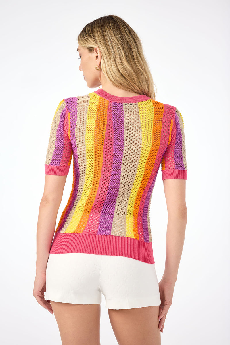 A woman faces away, wearing the Marbella Short-Sleeve Pullover— a sheer knit top with vertical pink, purple, orange, and yellow stripes—paired with white shorts. The background is plain white.