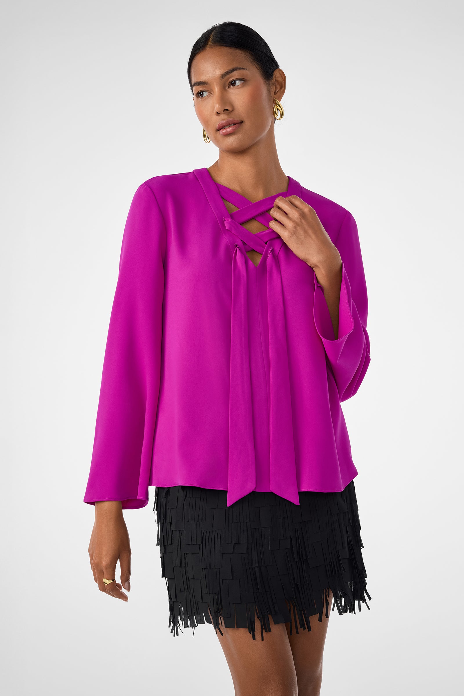 A woman wears the Picabella Top, a bright fuchsia long-sleeve blouse with crisscross neckline detail, paired with a short black fringe skirt. She accessorizes with gold hoop earrings and has her hair styled back.