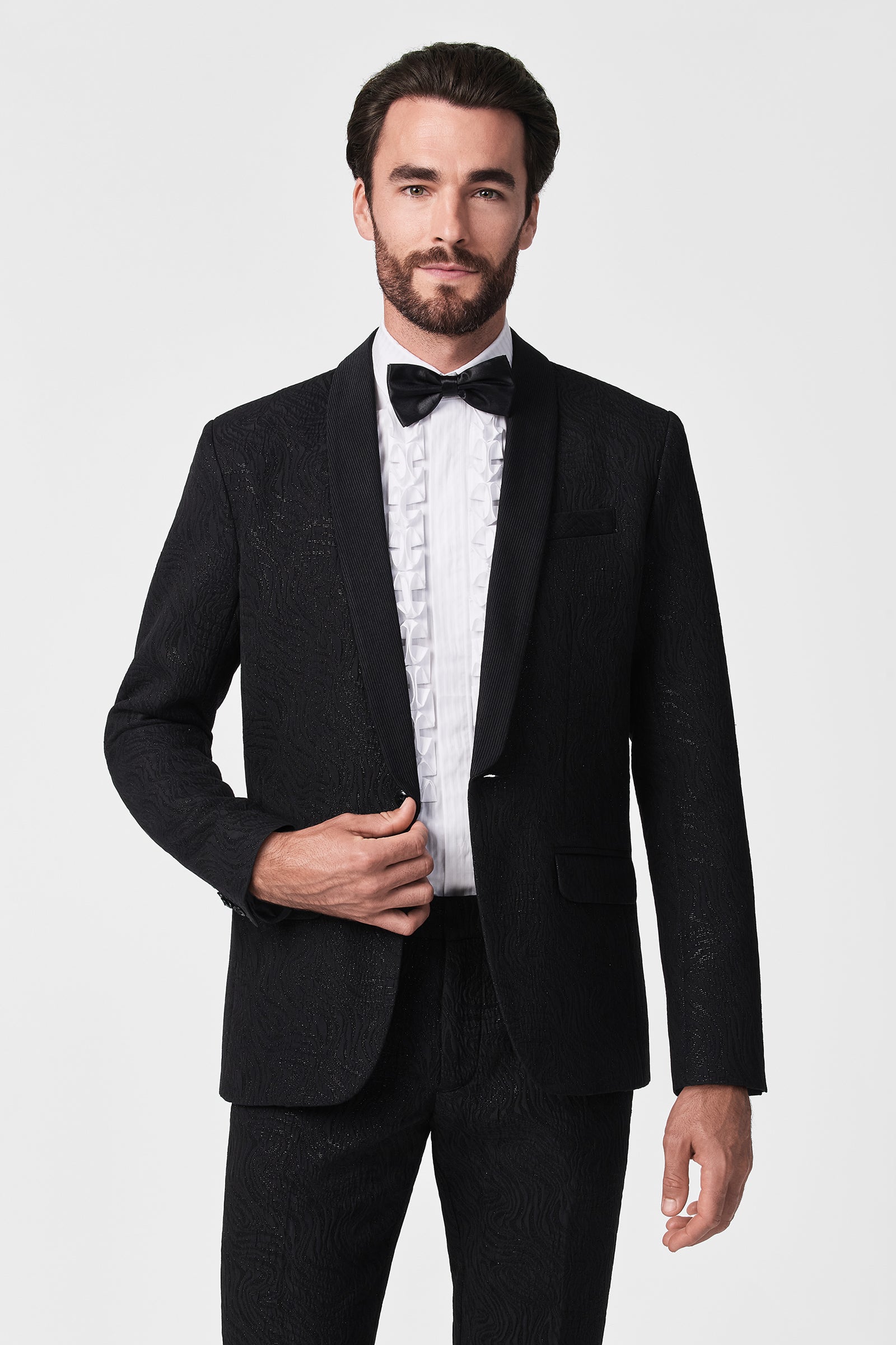 A man with dark hair and beard wears the Thurston Blazer, a black tuxedo jacket, paired with a pleated white dress shirt and black bow tie, standing against a plain white background.