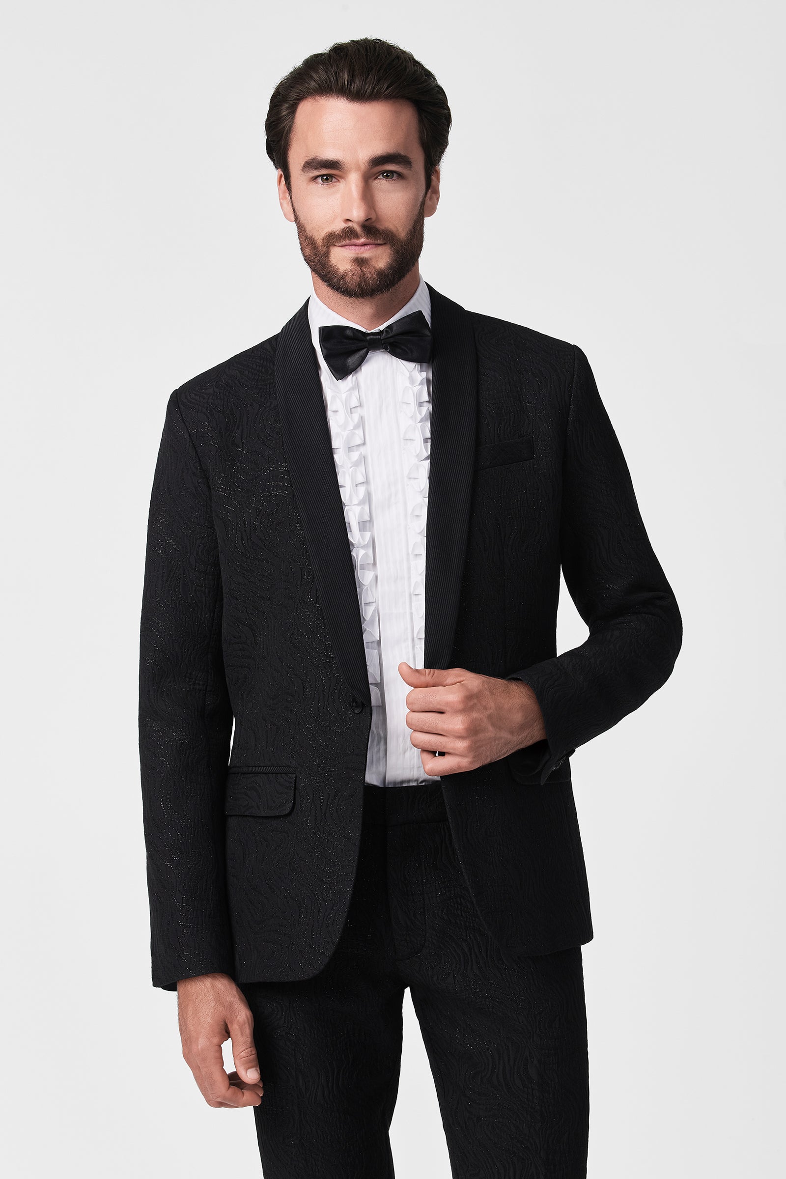 A man with dark hair and a beard wears the Adrian Tux Trouser, paired with a black shawl-collar tuxedo jacket, white dress shirt, and black bow tie. He stands against a white background, facing forward and adjusting his jacket.