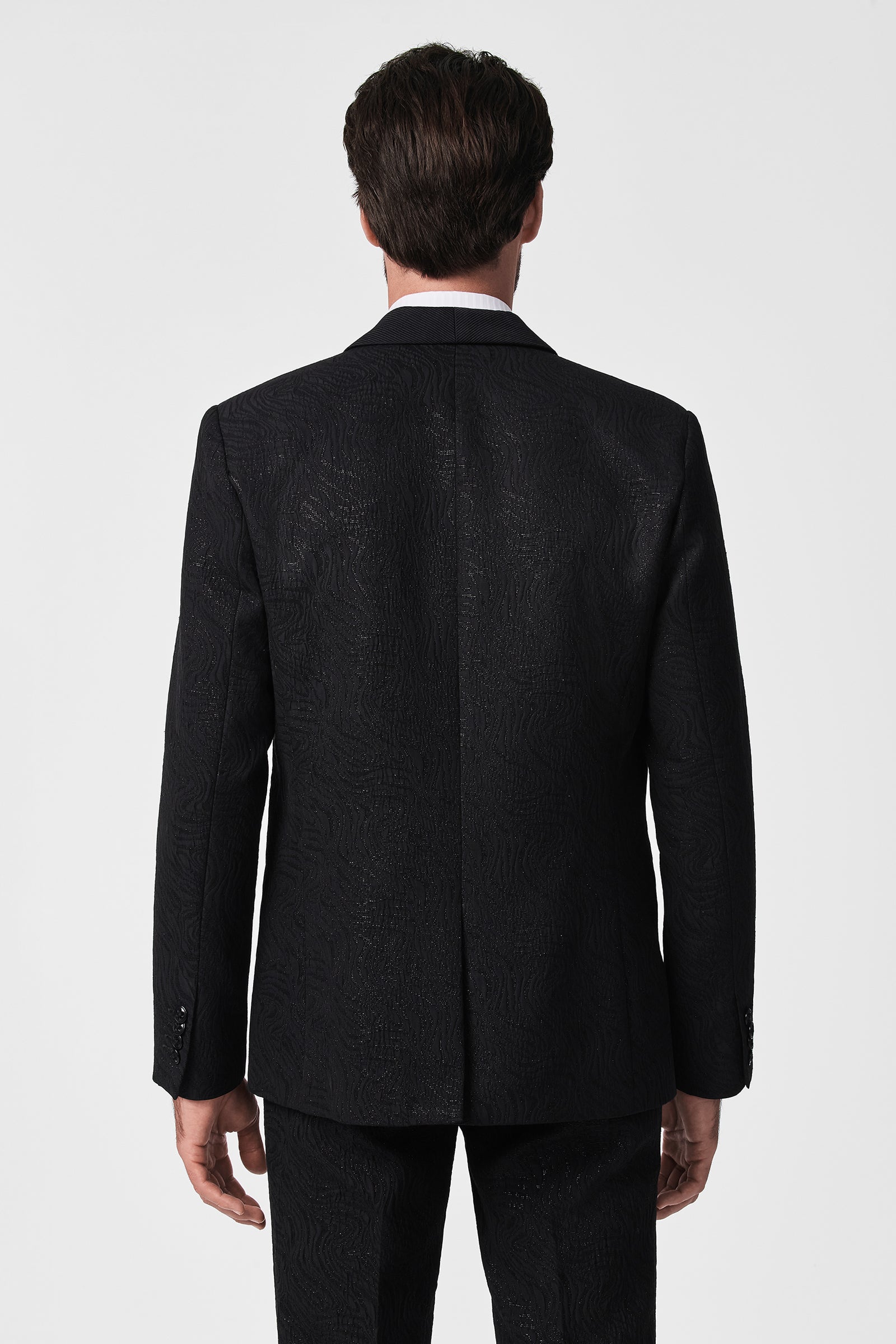 A man is seen from behind wearing the Adrian Tux Trouser, a textured black suit pant, standing against a white background with relaxed arms and neatly styled dark hair.