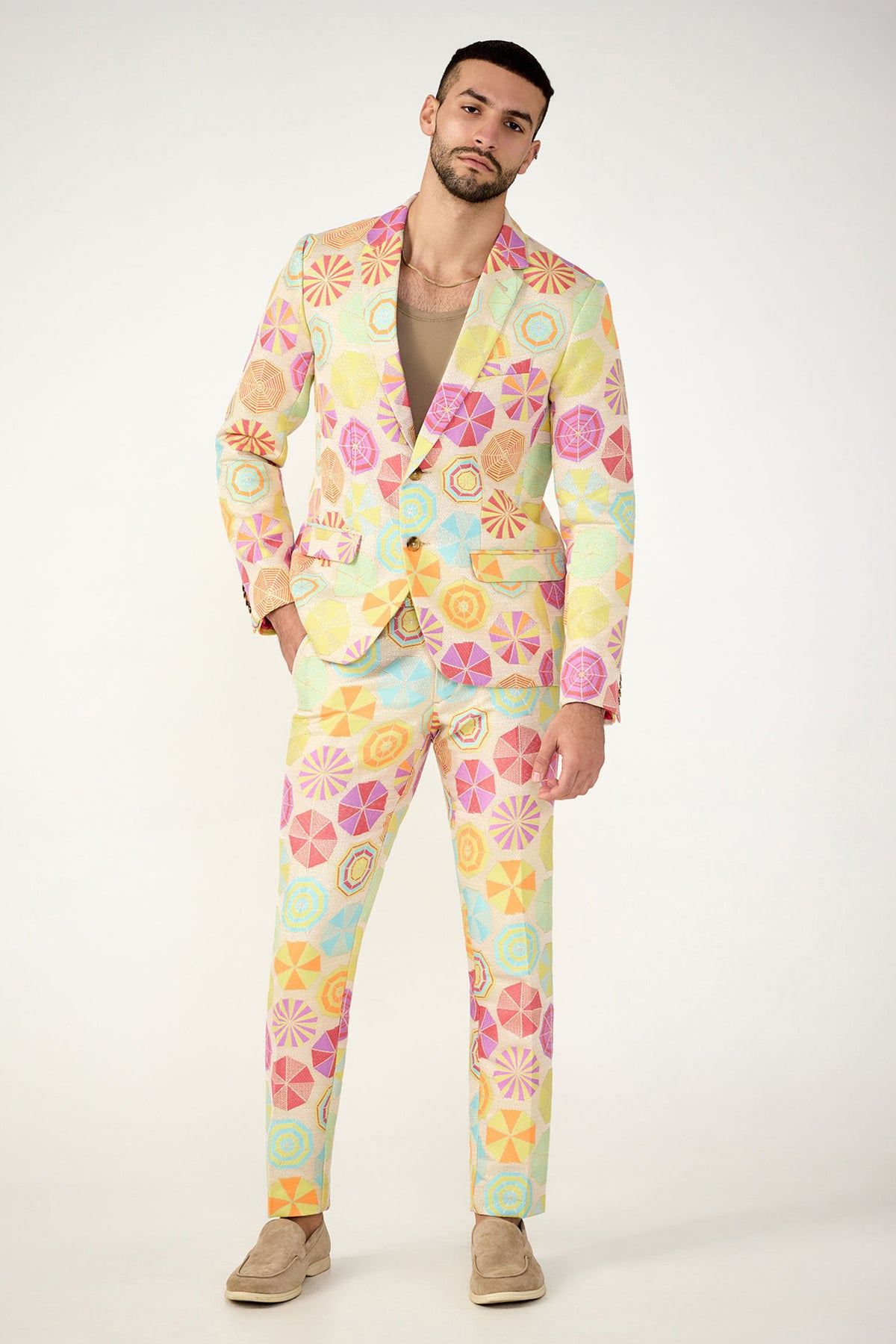 A man models the Clyde Slim Trouser - Parapluie Jacquard, featuring a geometric pastel pattern in yellow, pink, orange, and turquoise. He pairs it with a beige shirt and tan slip-ons against a plain white background.