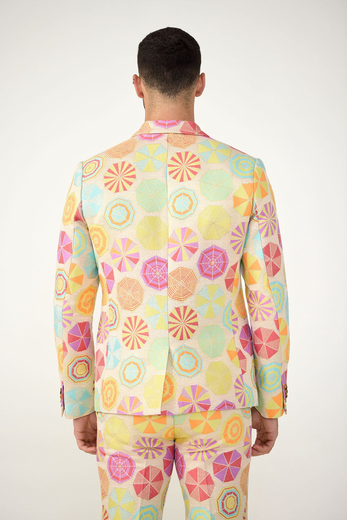 A person stands facing away, wearing the Thurston Blazer - Parapluie Jacquard, a colorful suit featuring a geometric pattern of circles and polygons in pastel pink, yellow, blue, and orange against a plain white background.