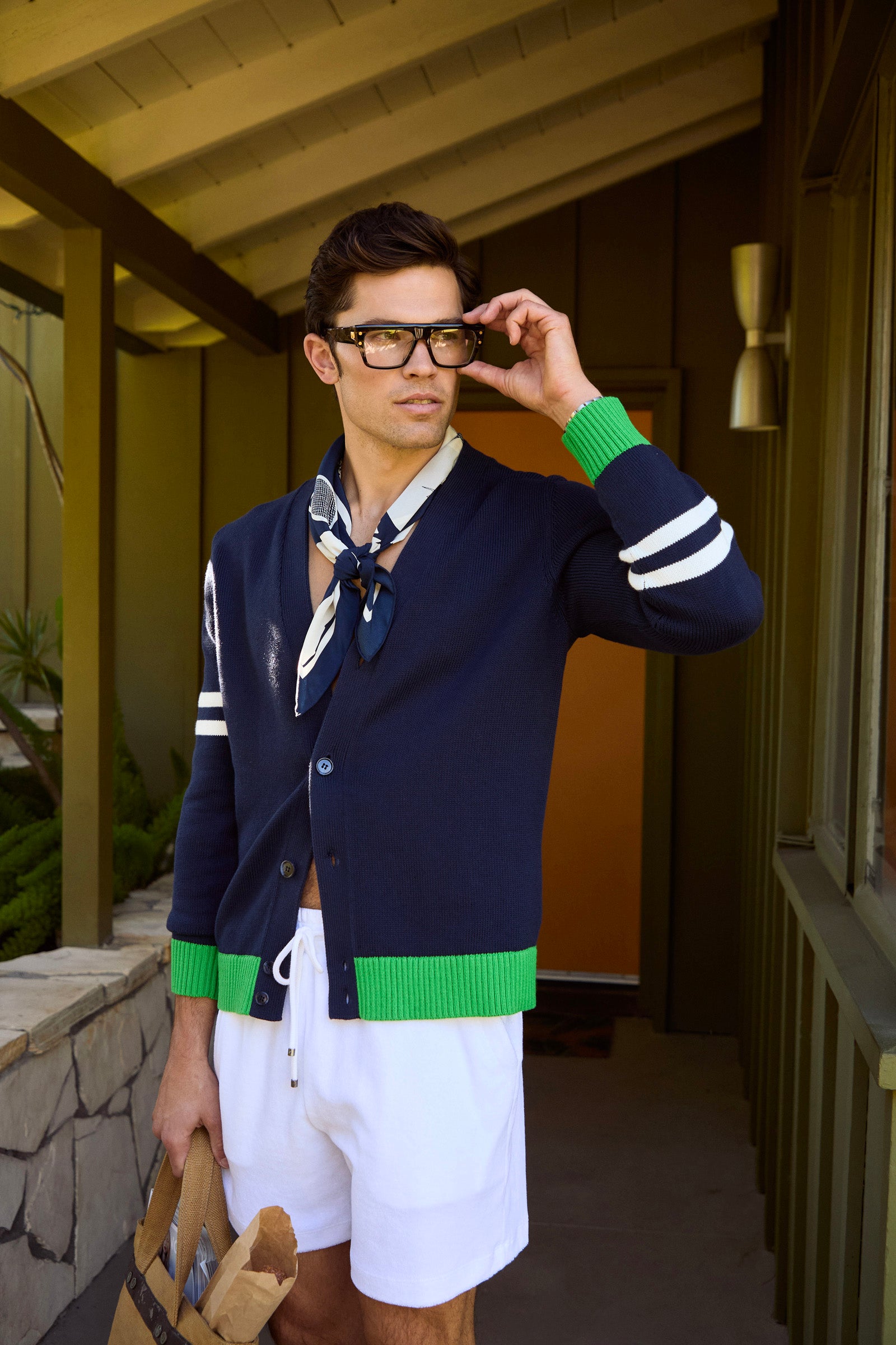 A man in the Amadeo Cardigan, featuring navy blue fabric and green cuffs, stands outside a house with a brown paper bag, wearing white shorts, glasses, and a neck scarf while gazing thoughtfully into the distance.