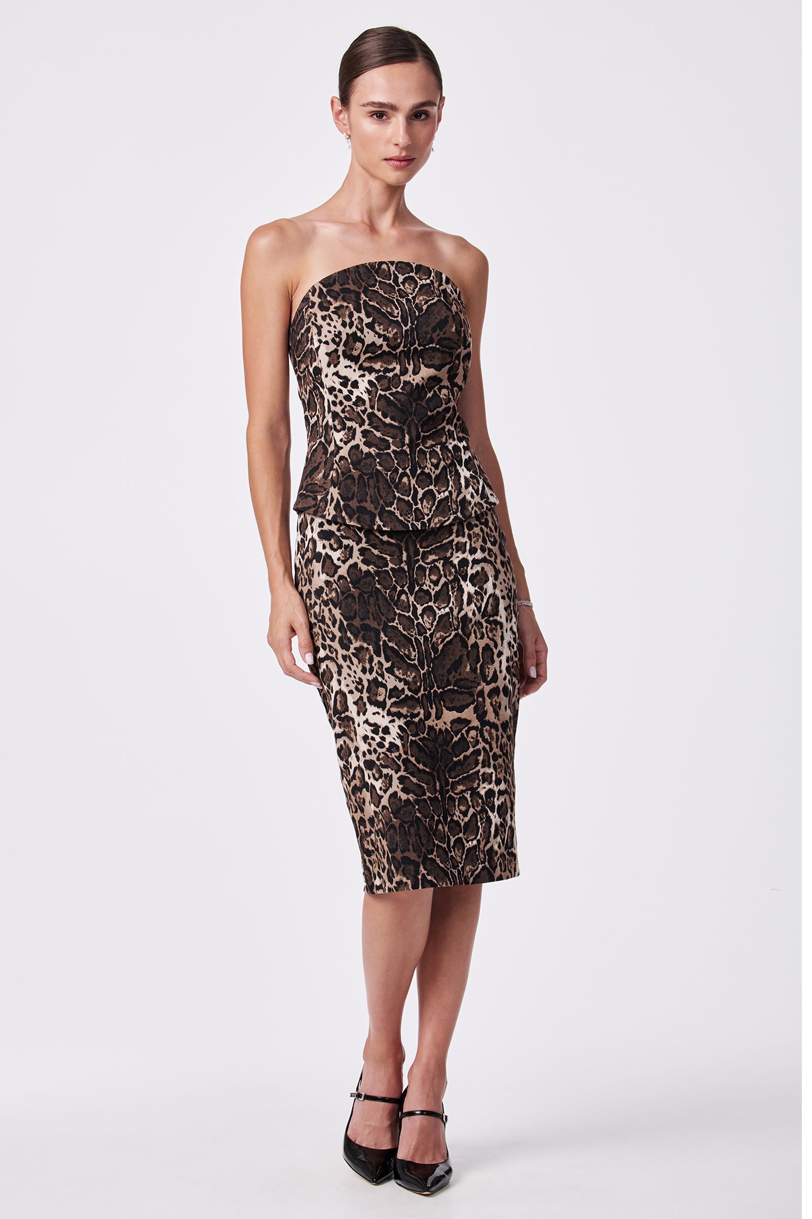A woman models the Osa Pencil Skirt, a fitted, knee-length skirt with a bold brown and black leopard print. She pairs it with her hair styled back and black pointed high heels.