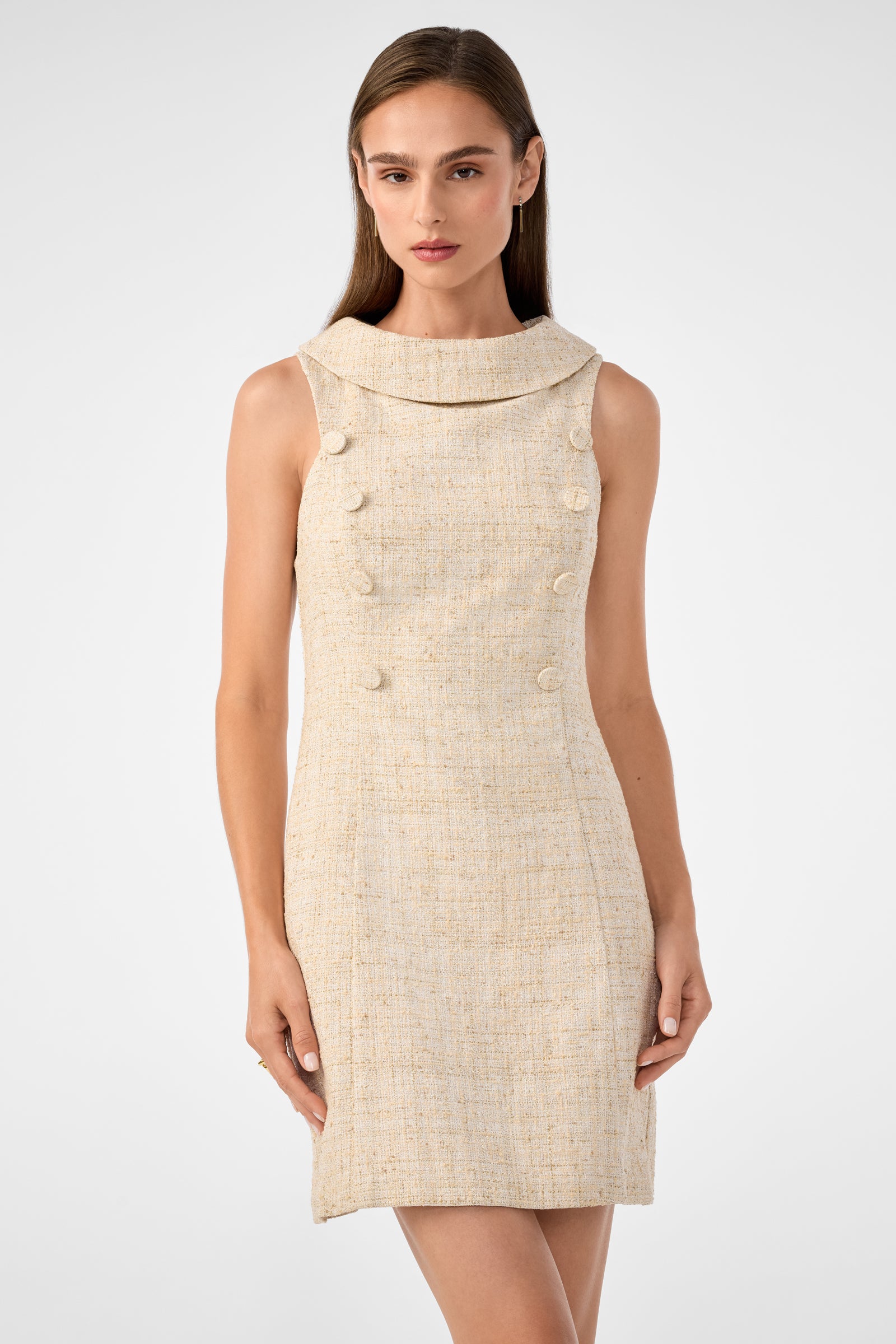A woman with straight brown hair models the Beziers Dress, a sleeveless beige tweed style with a wide collar and front decorative buttons, standing against a plain light gray background.