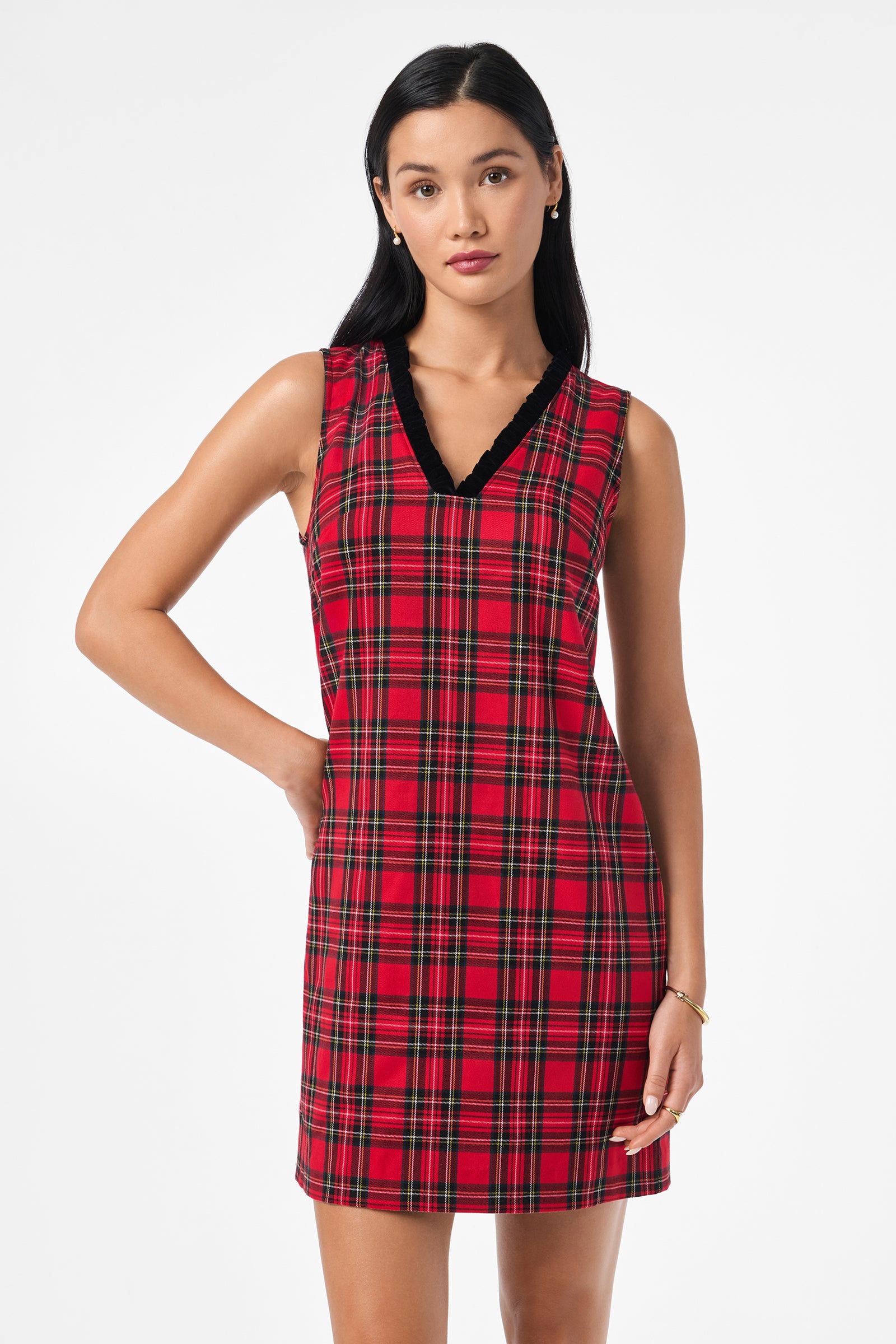 A woman with long black hair models the Revah Dress—a sleeveless red plaid style with black trim—posing with one hand on her hip against a plain white background.