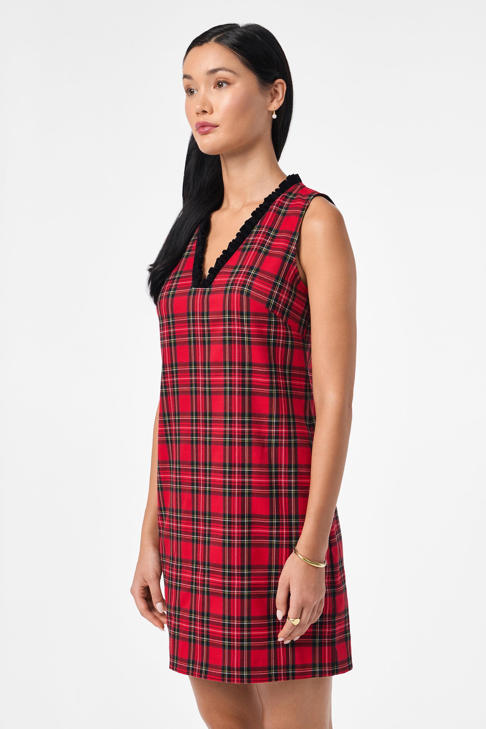 A woman with long dark hair wears the Revah Dress—a sleeveless red plaid style with a black scalloped V-neckline—as she stands against a plain white background, gazing slightly to the side.
