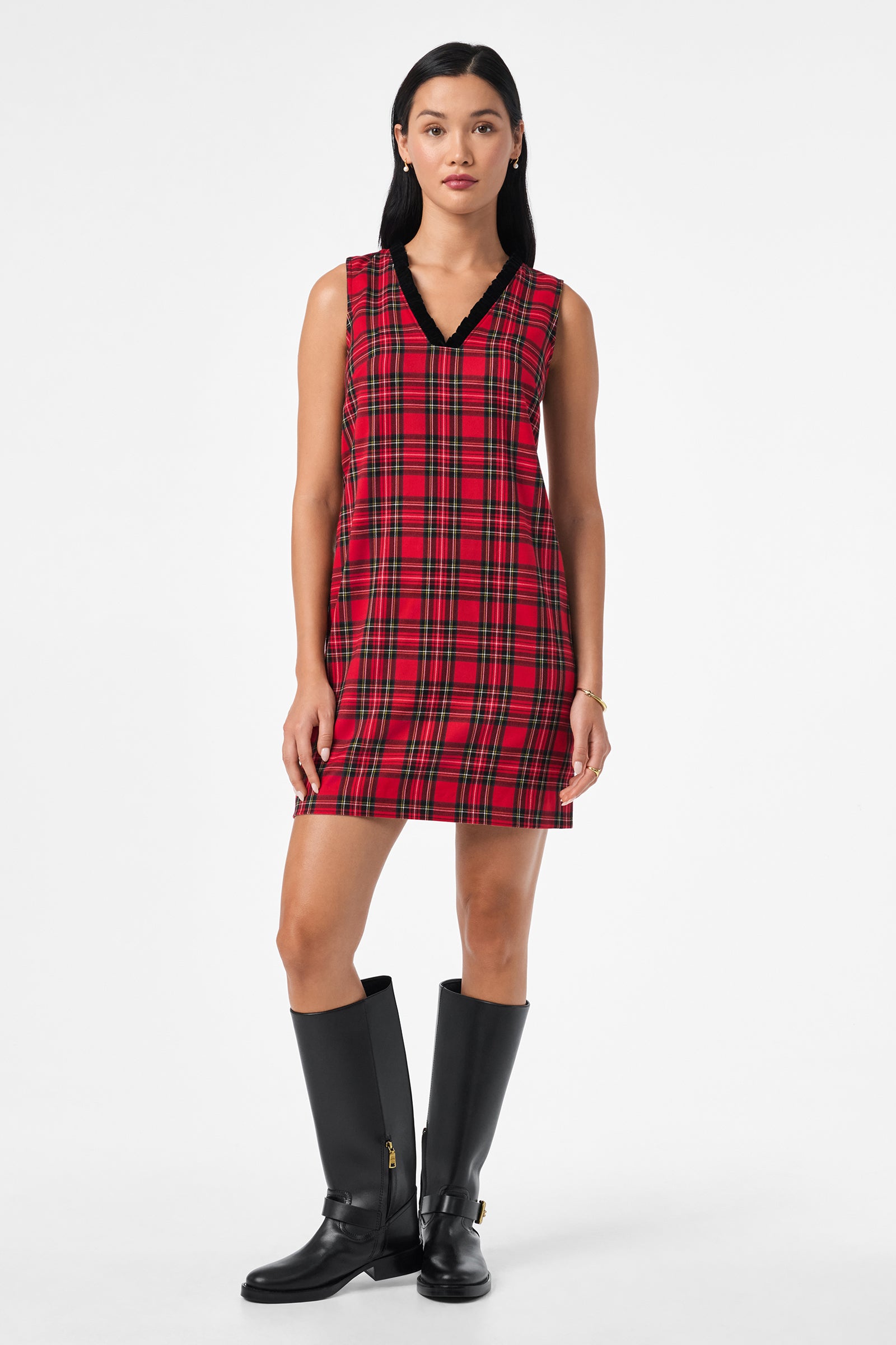 A woman stands against a plain background wearing the Revah Dress, a sleeveless red plaid dress with a black V-neck trim, paired with black knee-high boots.