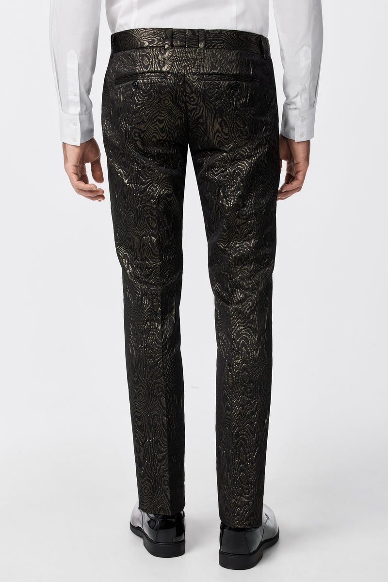 A person is seen from the back wearing the Clyde Slim Trouser - Fette Swirl, featuring a shiny gold pattern, belt loops, and pockets, paired with a white dress shirt and black dress shoes.