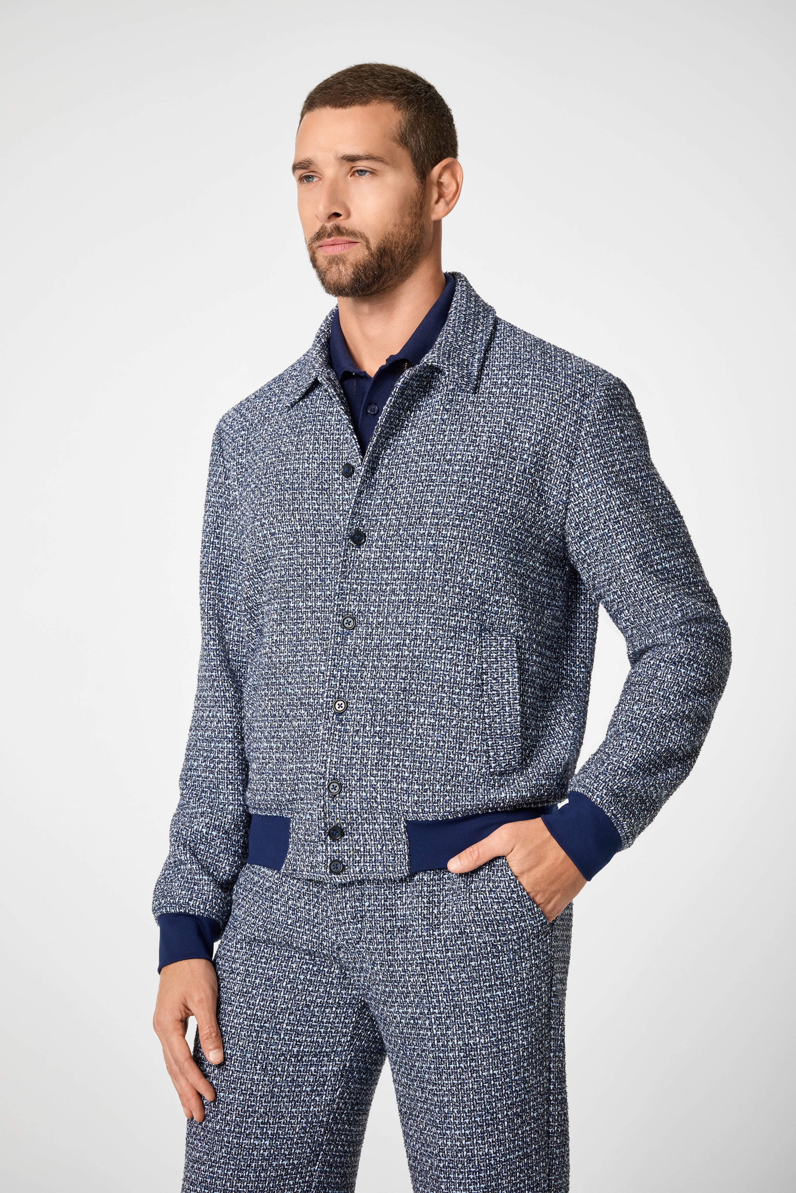 A man with short brown hair and a beard wears the Lowen Jacket, featuring a textured blue and white design, with his left hand in his pocket, standing against a plain light background.