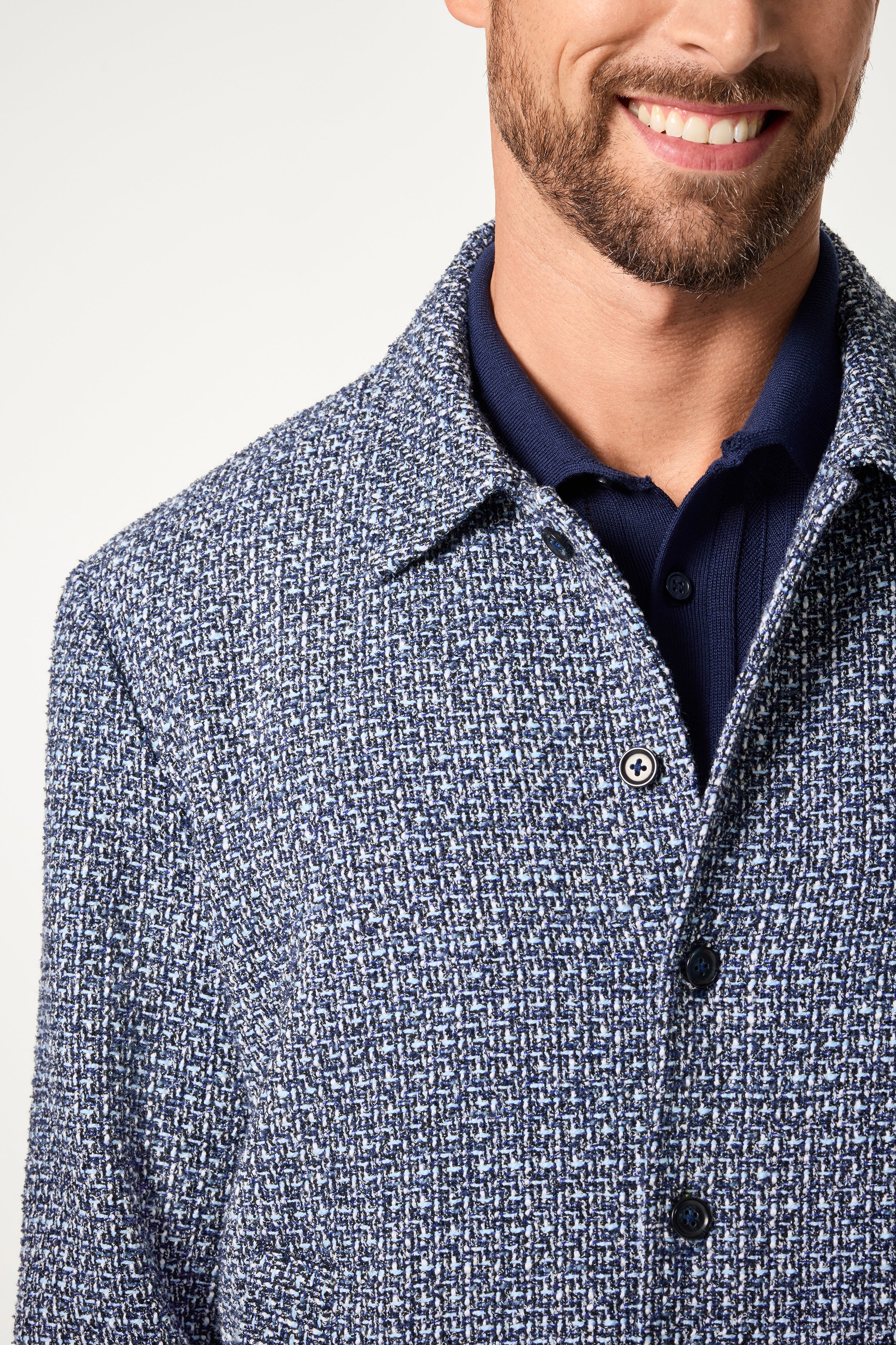 A close-up of a smiling man wearing the Lowen Jacket, a textured blue and white piece layered over a dark blue collared shirt, shown from the shoulders up against a plain light background.