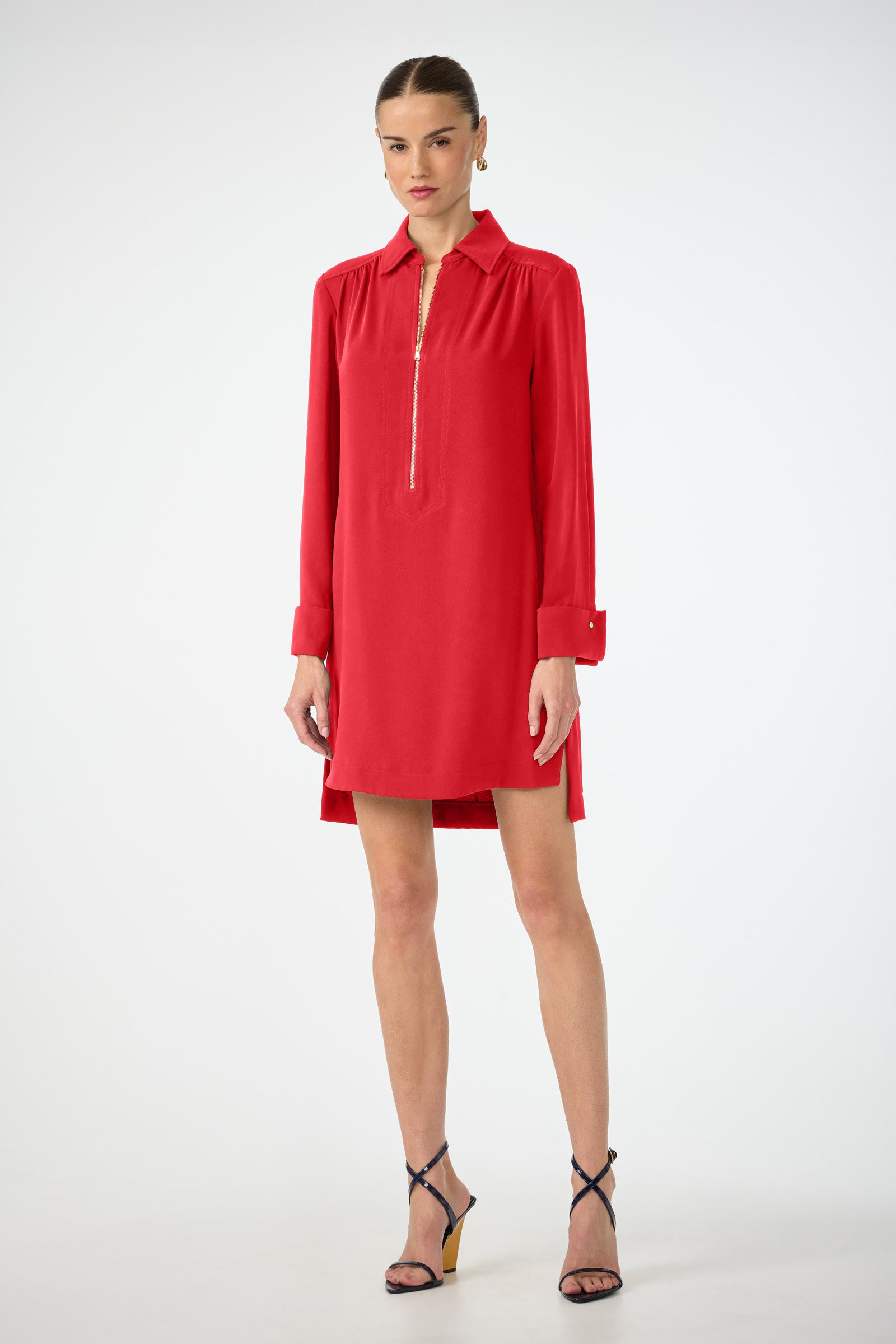 A woman stands against a white background wearing the Darya Long-Sleeve Dress—a red collared dress with a front zipper and high-low hem—paired with black strappy heels.