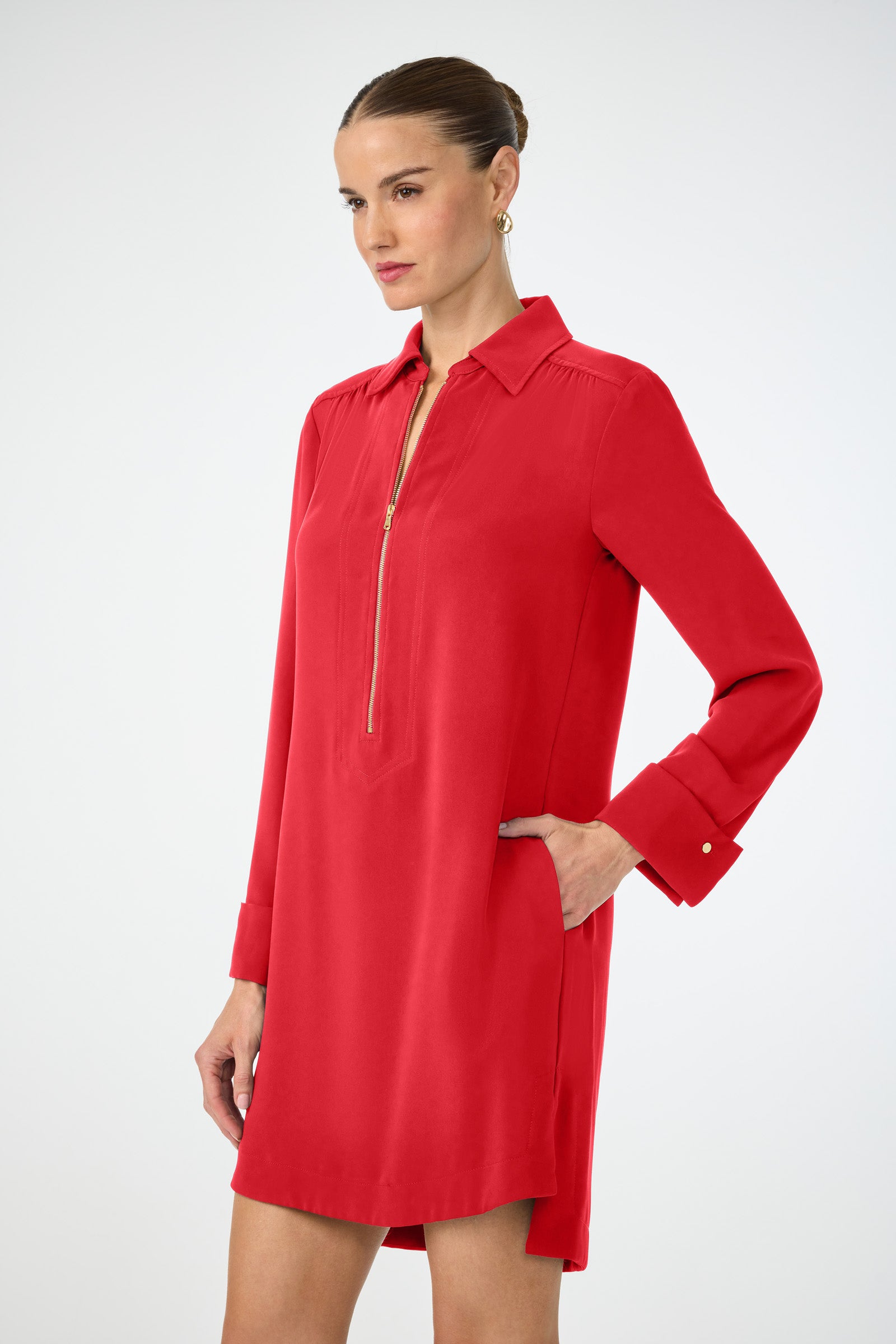 A woman stands against a white background wearing the Darya Long-Sleeve Dress in bright red, featuring a collared neckline, front zipper, and side pockets. She has her hair pulled back and wears gold hoop earrings.