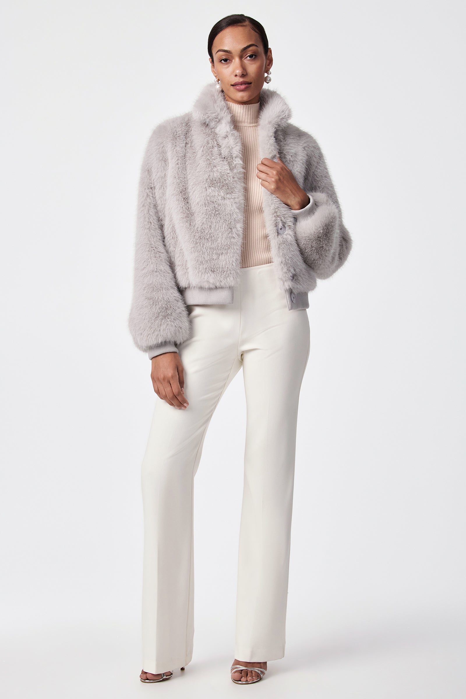 A woman stands against a plain background wearing the Morris Short Faux Fur Coat in light grey, a beige ribbed turtleneck, white high-waisted pants, and beige open-toe heeled sandals.