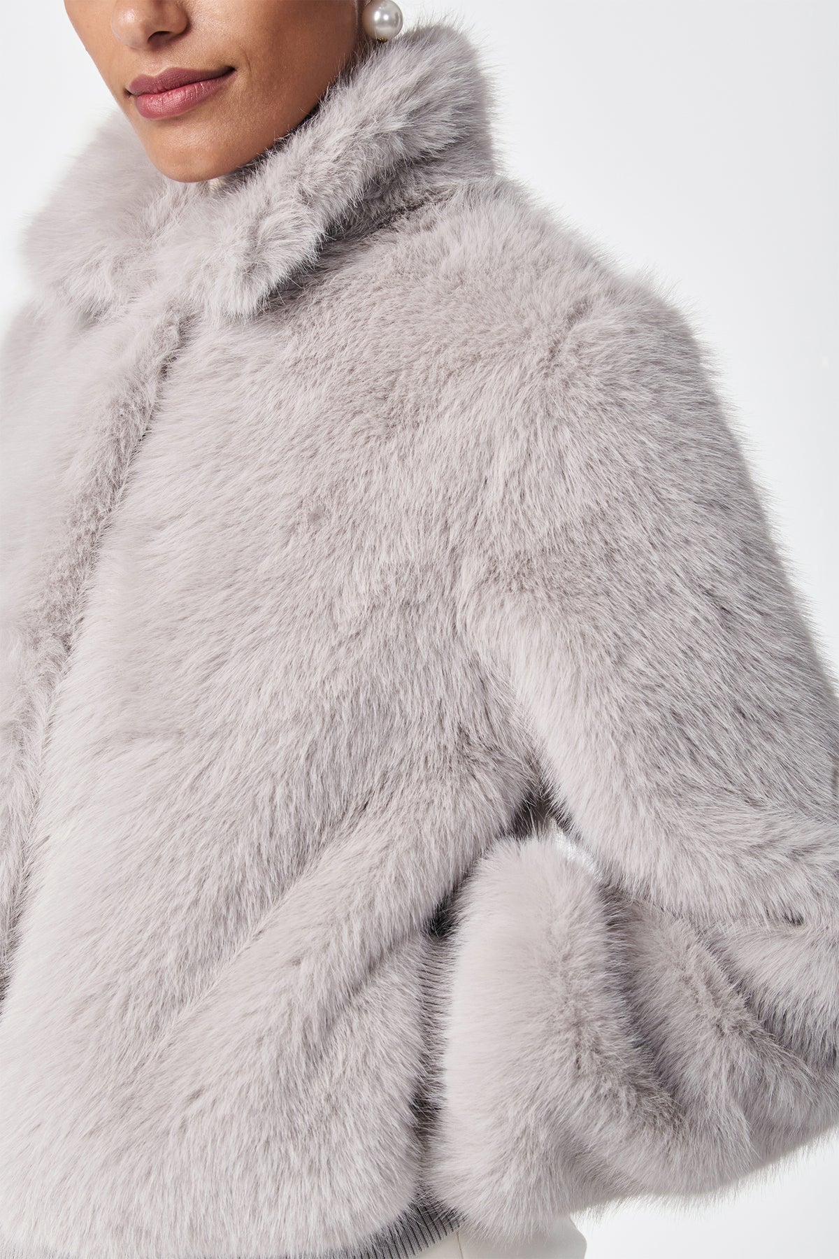 A woman models the Morris Short Faux Fur Coat in light gray, featuring a high collar and soft texture, with one hand in her pocket and a visible pearl earring against a plain white background.