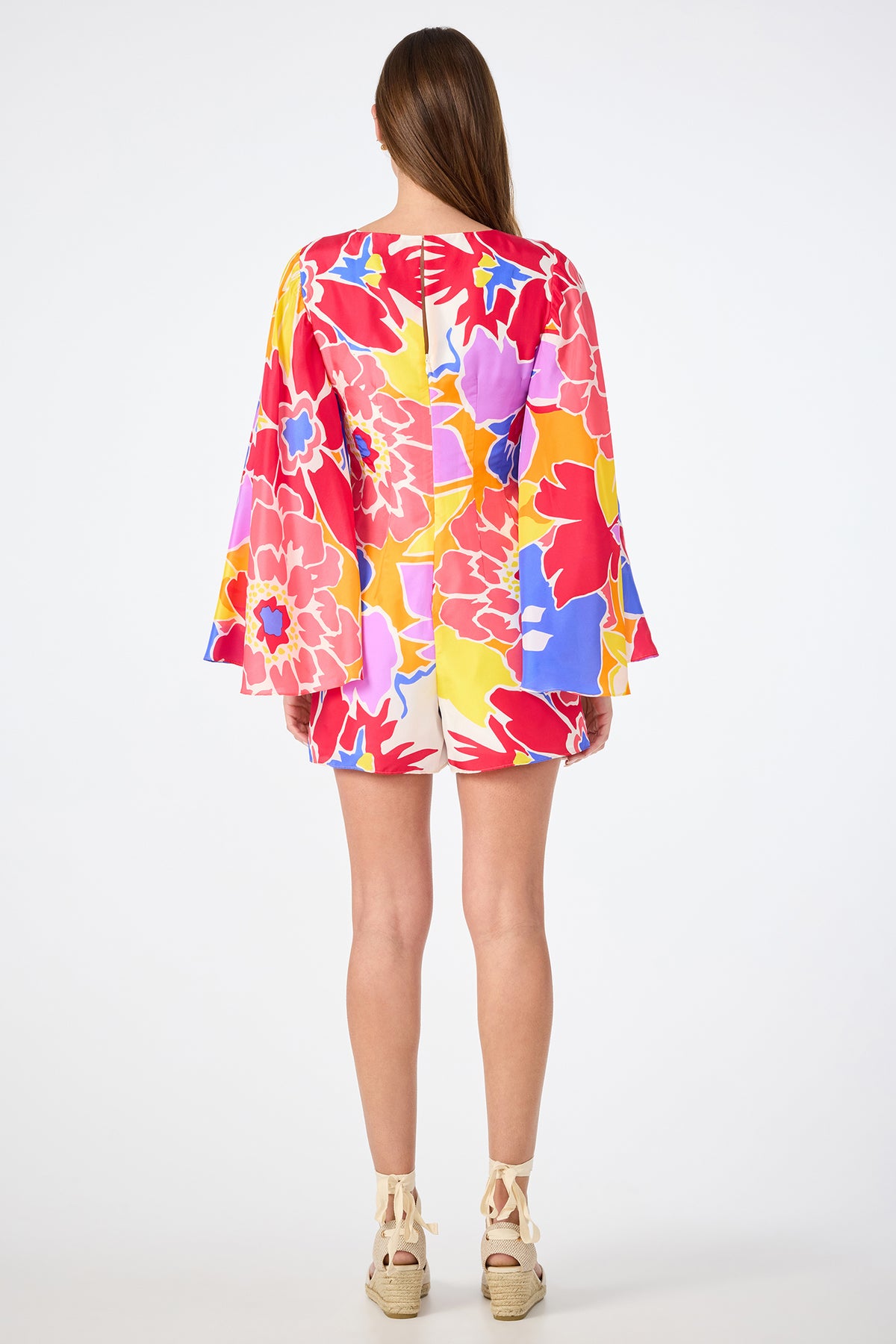 A woman with long brown hair faces away, wearing the Miera V-Neck Romper - Soie Paradis, a bright floral-print romper with long sleeves and multicolored flowers, paired with beige wedge sandals.