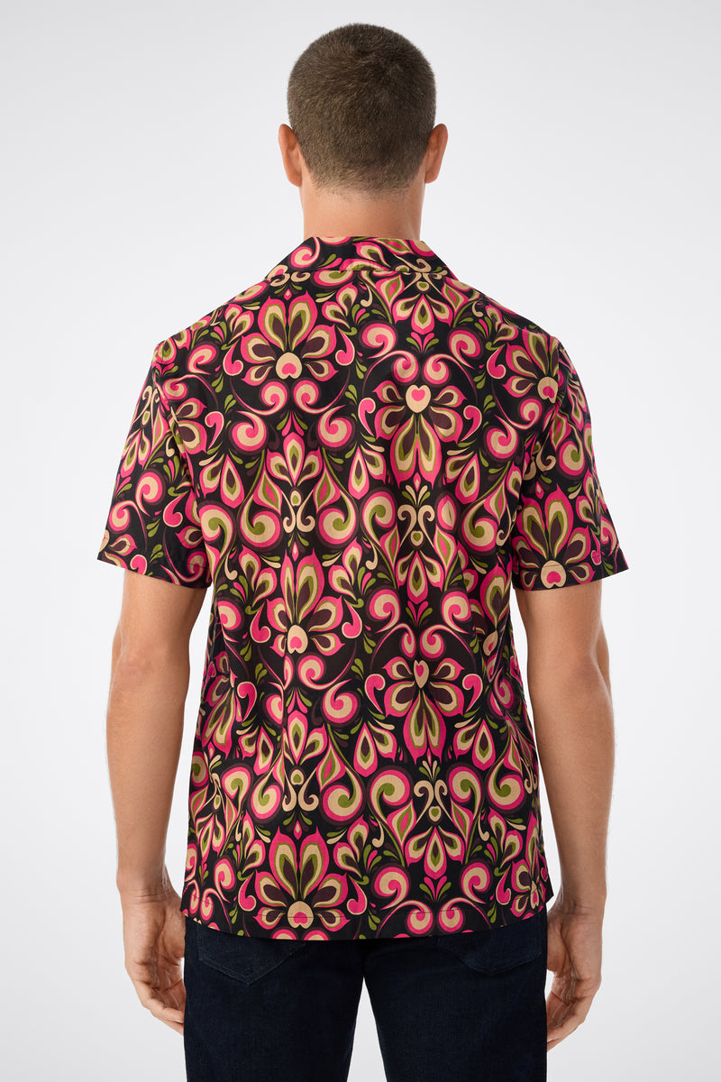 A person with short hair is shown from behind wearing the Felix Camp Shirt - Sanctuary, which features a bold swirling pink, green, and black pattern, along with dark pants.
