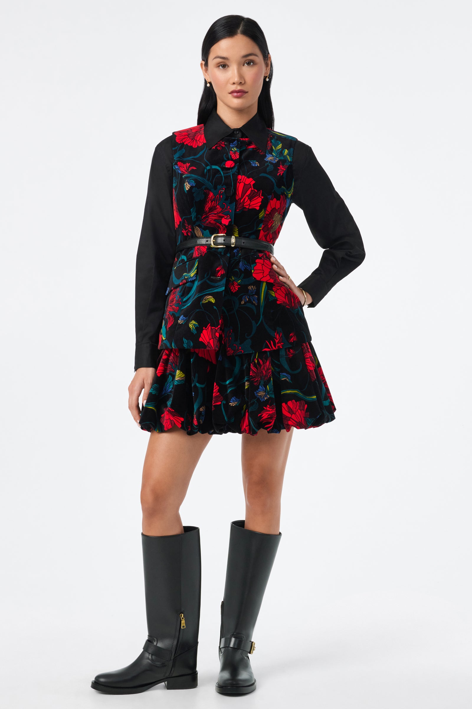 A person stands facing forward, wearing the Kary Top—a black collared shirt—under a sleeveless dress with a bold red and green floral pattern, belted at the waist, and paired with black knee-high boots.