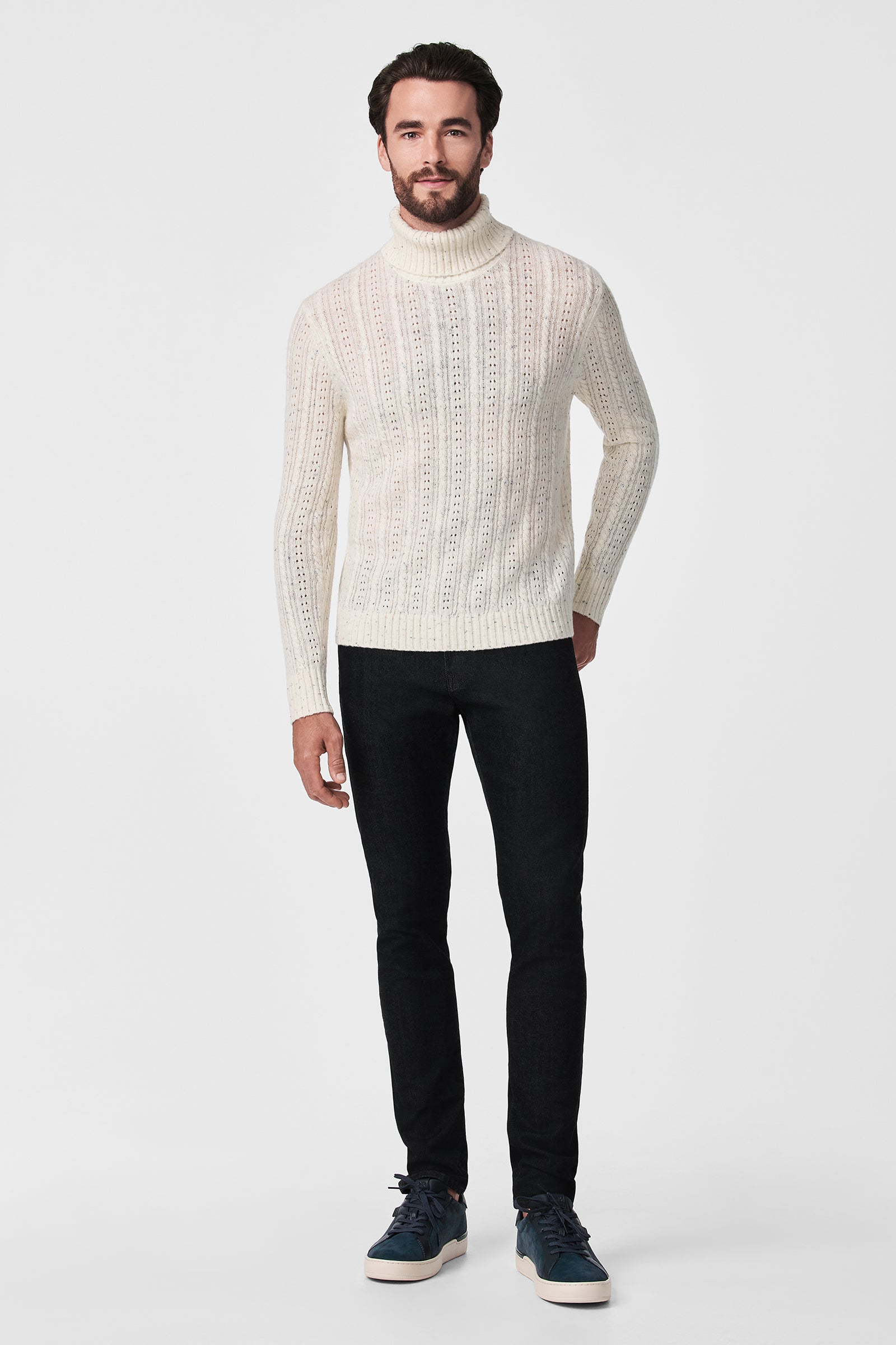 A man stands against a white background wearing the cream Wesley T-Neck sweater, black slim-fit pants, and navy blue sneakers.