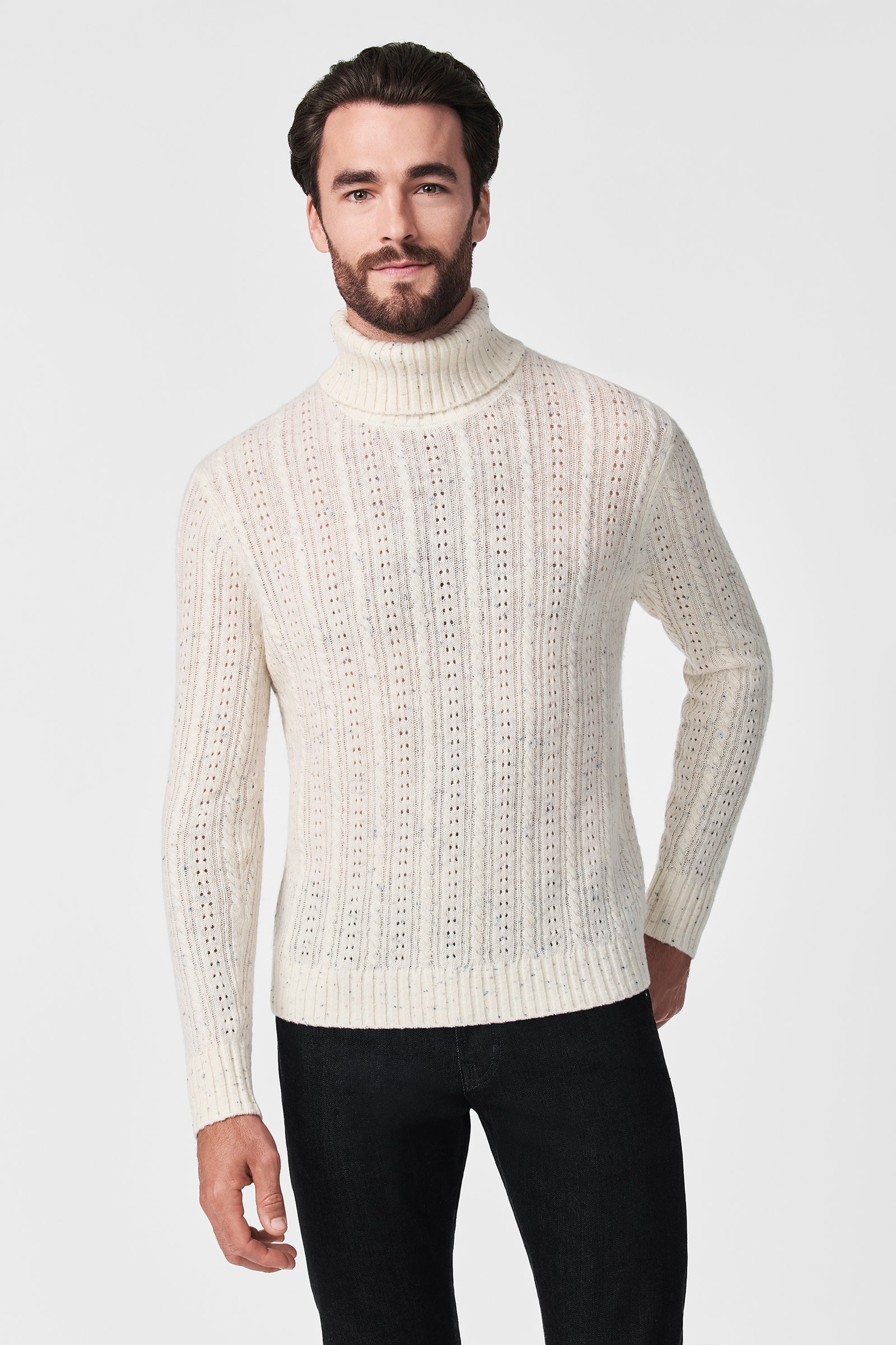 A man with dark hair and a beard wears the Wesley T-Neck, a white cable-knit turtleneck sweater, paired with black pants against a plain white background.