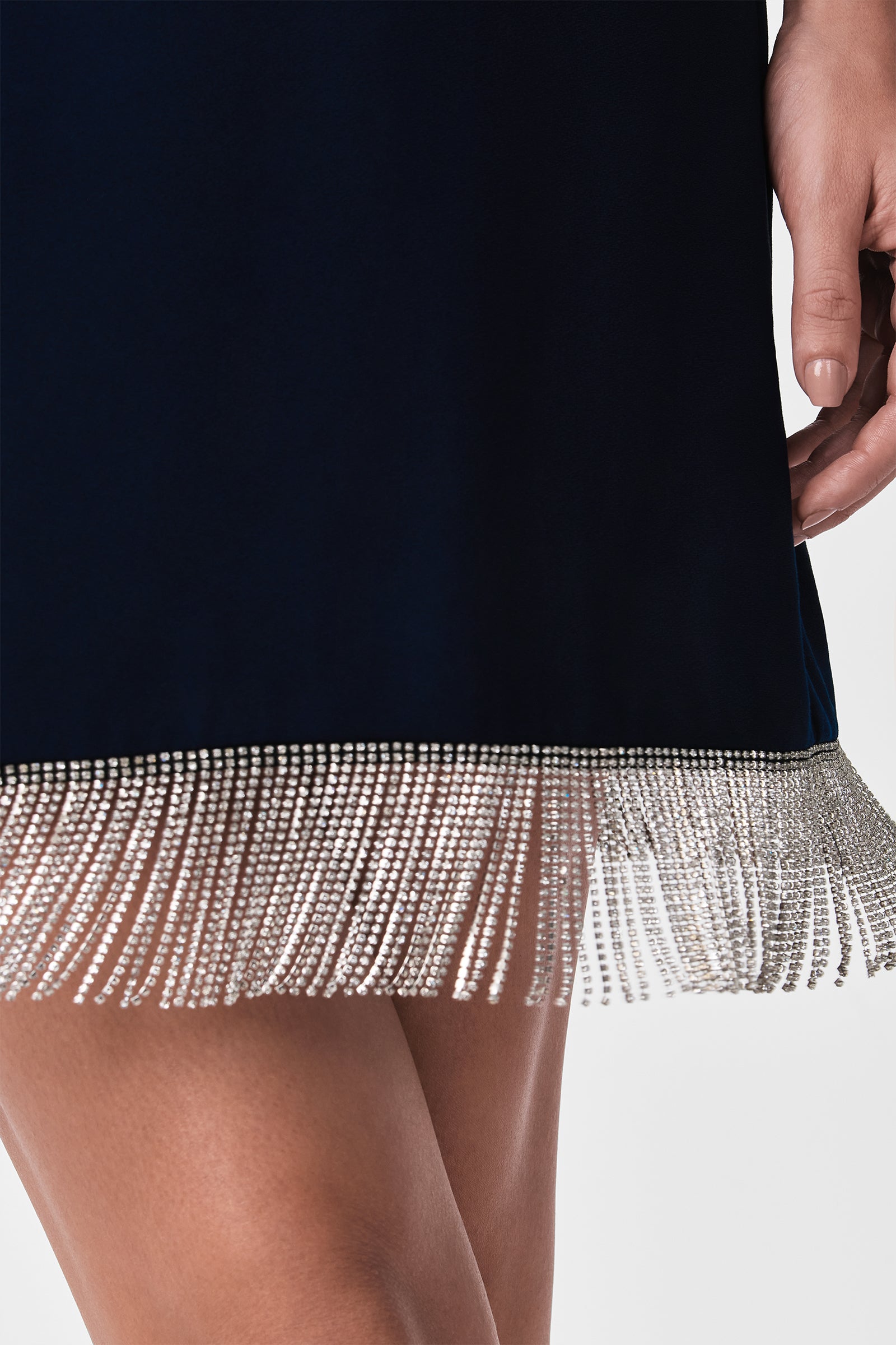 A close-up of a person wearing the Top Shelf Dress, featuring dark blue fabric with silver fringe along the hem. Their hand rests by their side, showing only part of the dress and legs.