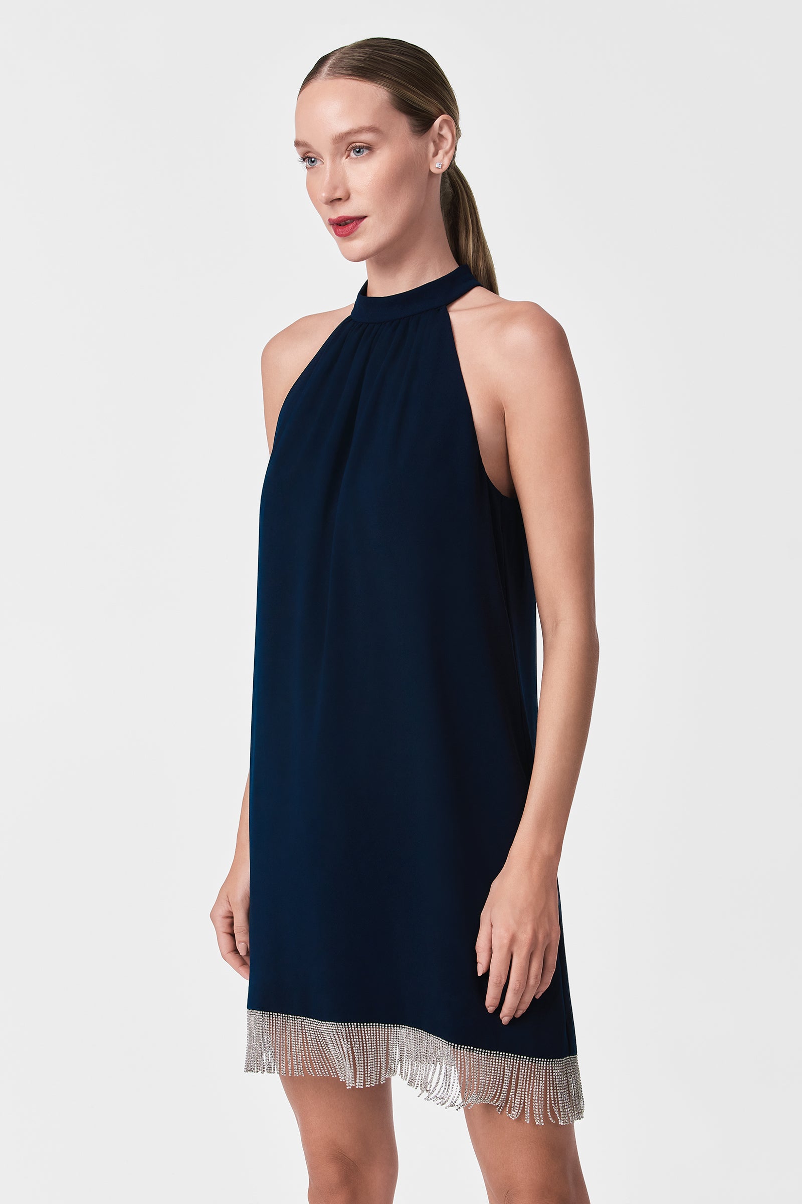A woman stands against a white background, her hair in a sleek ponytail, wearing the Top Shelf Dress—a sleeveless, high-neck navy blue style featuring silver fringe along the hem.