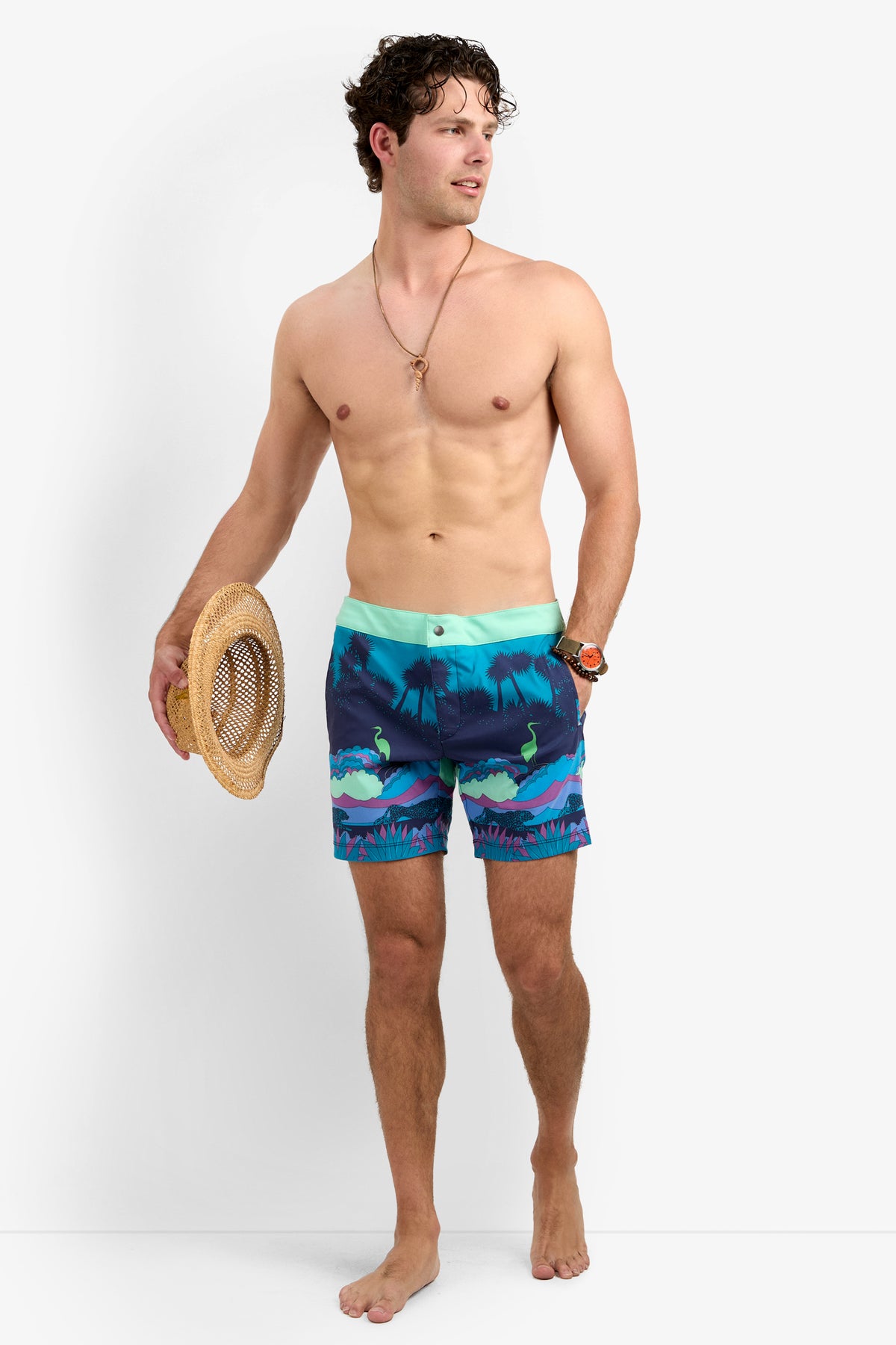 A shirtless man, barefoot against a plain white background, looks to the side while wearing the Honolulu Board Short - Jungle and holding a straw hat in one hand.