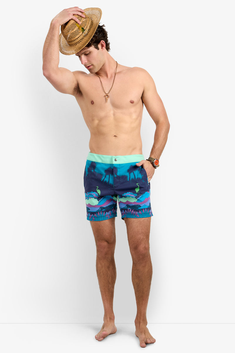 A shirtless man stands barefoot wearing the Honolulu Board Short - Jungle, featuring blue and green tropical prints. He holds a straw hat on his head and accessorizes with a necklace and wristwatch while looking down.