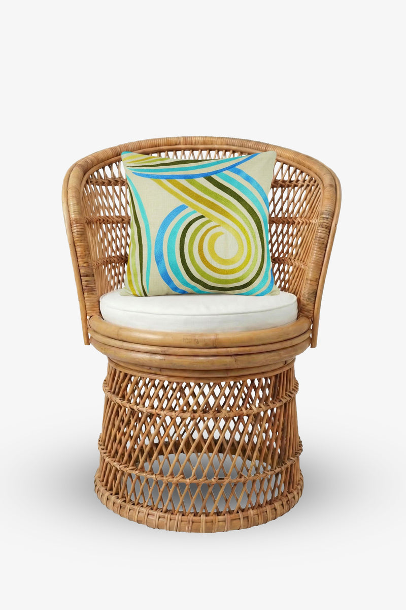 A round wicker chair with a beige seat cushion is styled with the Calexico Embroidered Square Pillow—measuring 20″x20″ and showcasing blue, green, and yellow swirled embroidery on a soft down-filled cover—set against a plain white background.