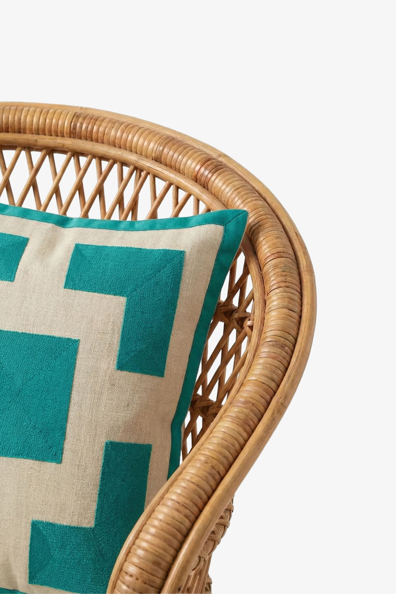 A close-up of a rattan chair featuring the Palm Springs Block Embroidered Square Pillow, showcasing its teal and beige geometric pattern inspired by Trina Turk, filled with plush down feathers, set against a plain white background.