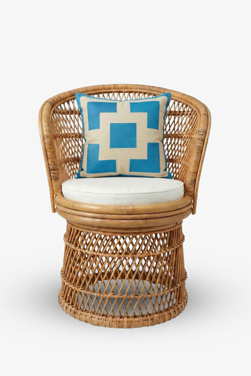 A round wicker chair with an open weave design and a beige seat cushion is accented by the Palm Springs Block Embroidered Square Pillow in blue and white geometric patterns, featuring a TRINA TURK HOME pillow cover with a plush down feather insert, placed on the backrest.