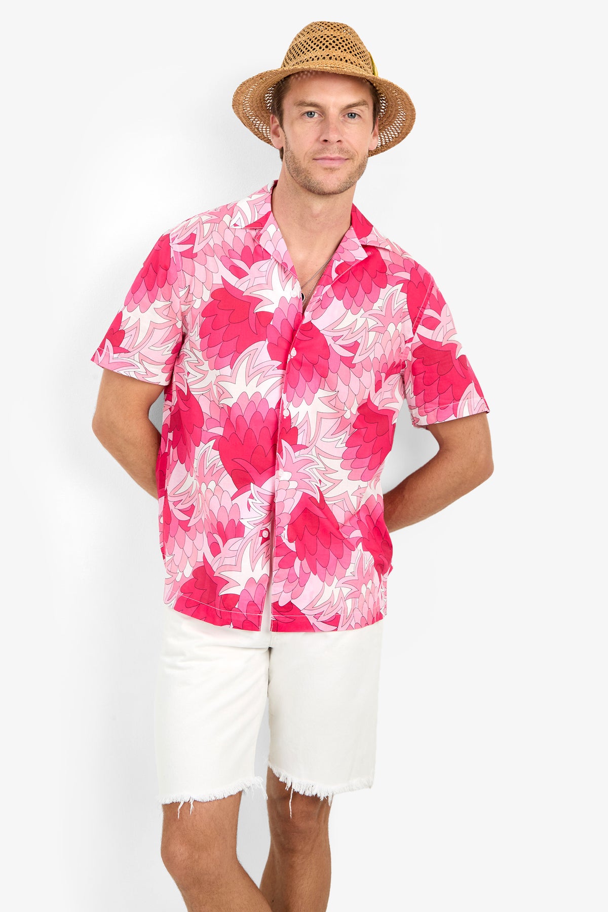 Man wearing the Felix Camp Shirt, featuring a pink floral print, pairs it with white shorts and a straw hat while standing against a light background, smiling with one hand behind his back.