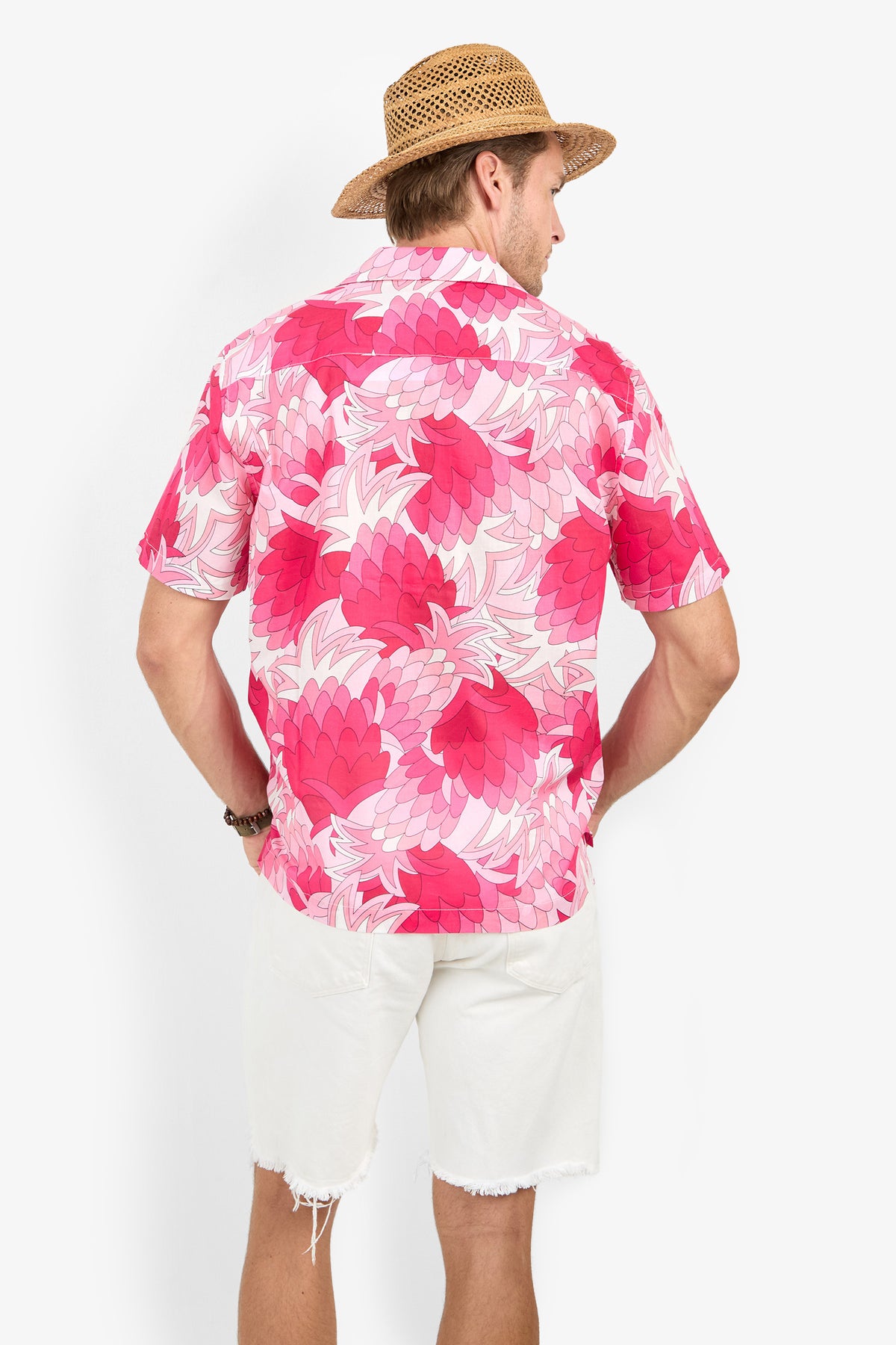 A man wearing a straw hat, Felix Camp Shirt in a pink pineapple print, and white shorts stands facing away from the camera against a plain white background.