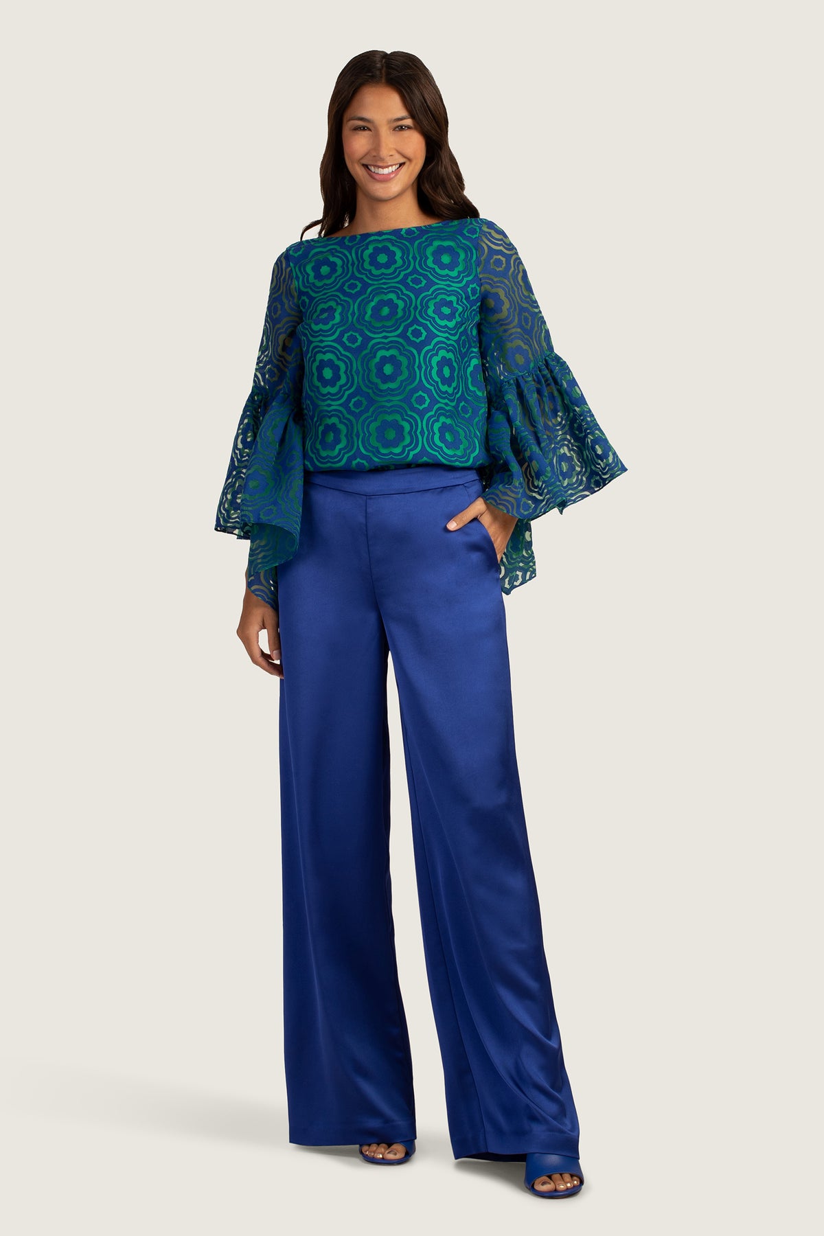 A woman smiles confidently in the Everly Top, a blue patterned blouse with flared, sheer sleeves, paired with high-waisted, wide-leg royal blue satin pants. Her long, dark hair frames her face as she poses with one hand in her pocket.