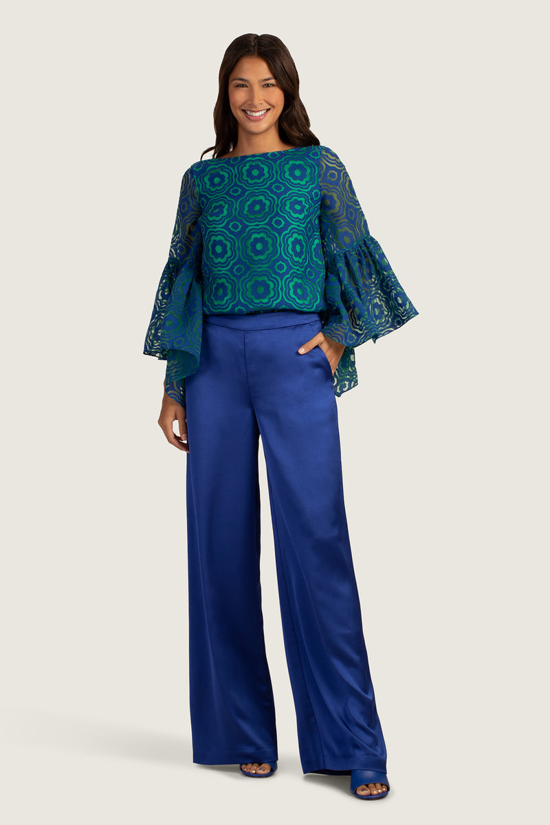 A woman smiles confidently in the Everly Top, a blue patterned blouse with flared, sheer sleeves, paired with high-waisted, wide-leg royal blue satin pants. Her long, dark hair frames her face as she poses with one hand in her pocket.