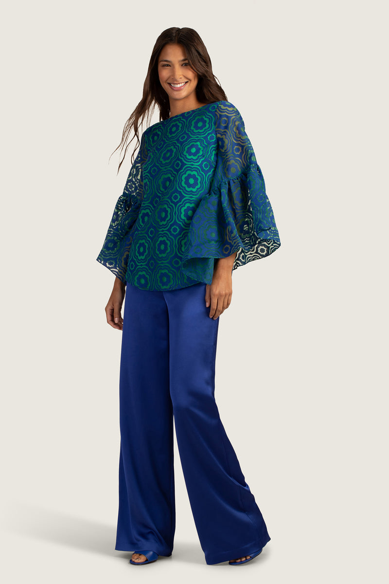 A woman smiles, wearing the Everly Top—a blue patterned special occasion blouse with flared sleeves—and wide-leg dark blue satin pants, standing against a light background.