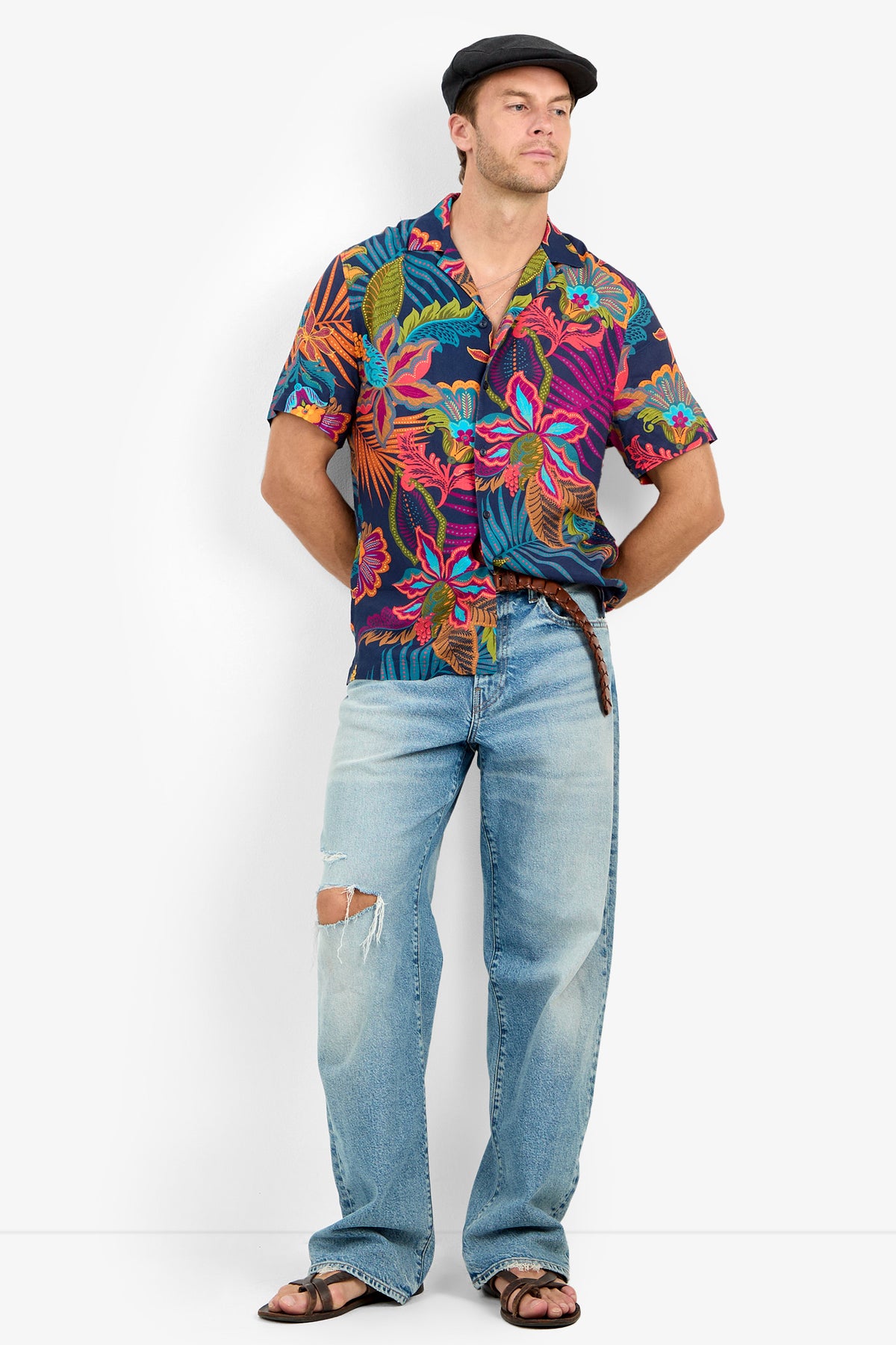 Man in a dark floral Felix Camp Shirt with a revere collar, light blue ripped jeans, brown sandals, and a black flat cap stands hands behind his back against a plain white background.