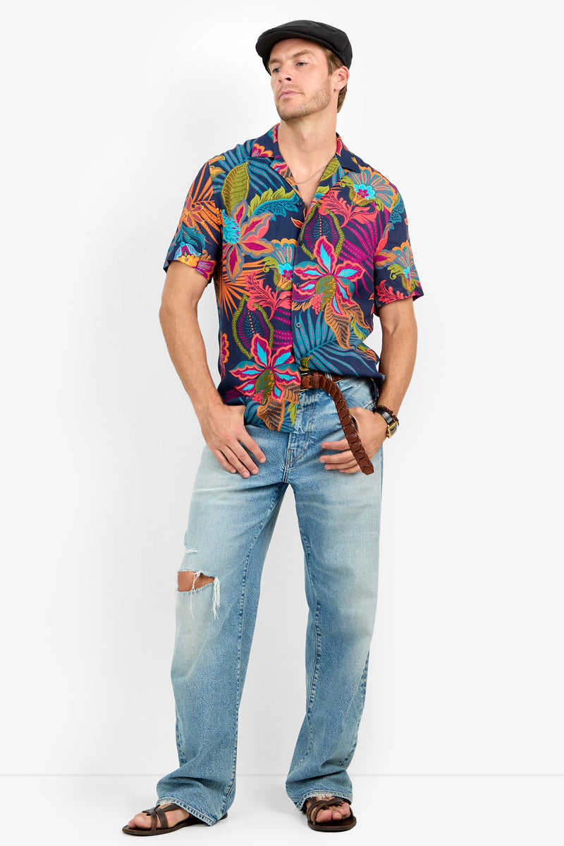 A man wearing a dark flat cap, Felix Camp Shirt with a summer print, ripped light blue jeans, brown belt, brown watch, and sandals stands with hands in pockets against a plain background.