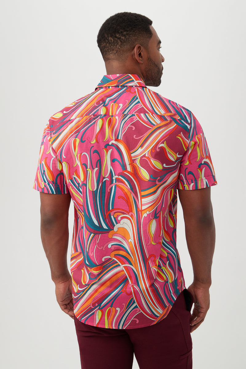 A man facing away wears the Slim Jim Shirt, a slim fit short-sleeve button-up featuring a bold swirling abstract pattern in pink, red, orange, yellow, blue, and white, paired with burgundy pants.