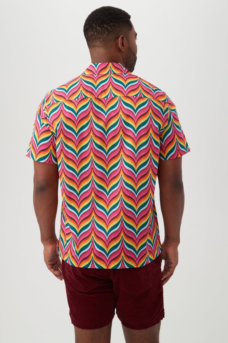 A man stands facing away, wearing the Felix Shirt—a colorful camp shirt with a revere collar and vibrant, wavy pink, yellow, green, and blue pattern—paired with burgundy shorts against a plain light background.