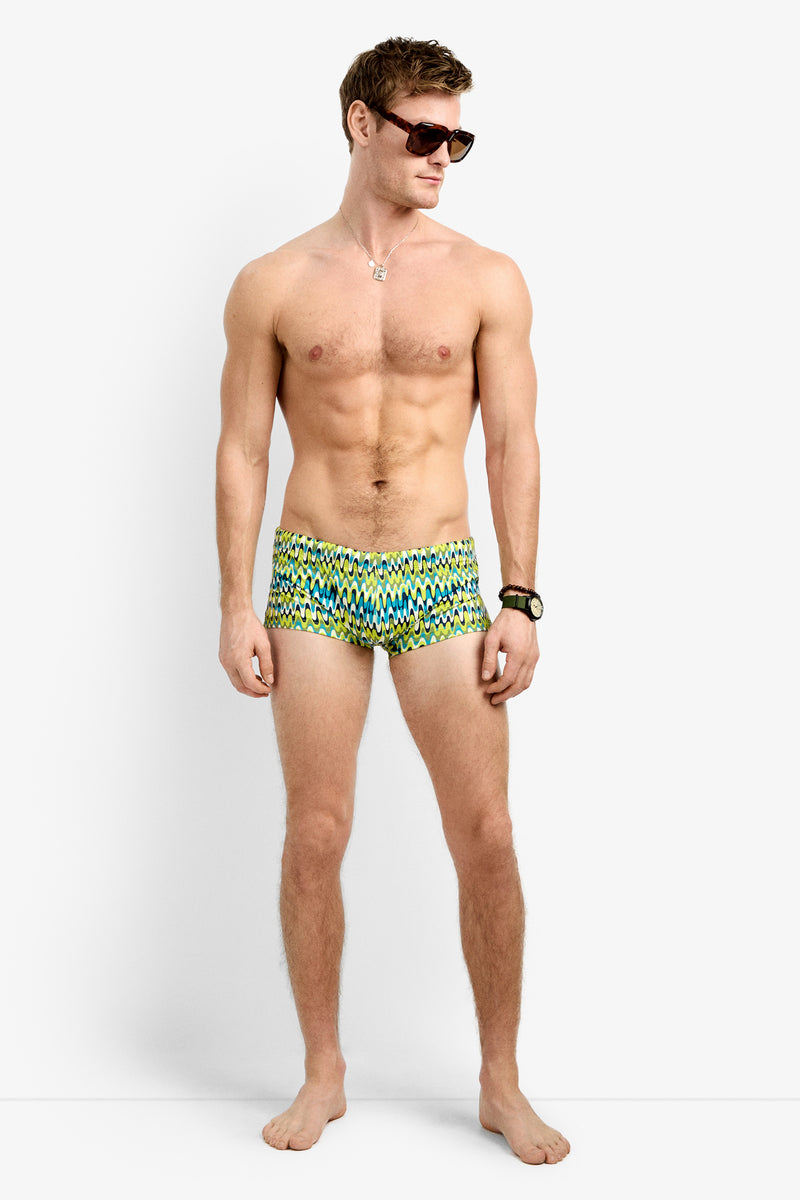 A shirtless man in sunglasses, a necklace, a wristwatch, and Sorrento Swim Trunk - Psychedelic Voyage stands barefoot against a plain white background.