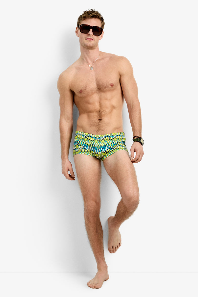 A shirtless man wearing the Sorrento Swim Trunk in Psychedelic Voyage, sunglasses, a necklace, and a watch poses barefoot with one leg bent against a white background.