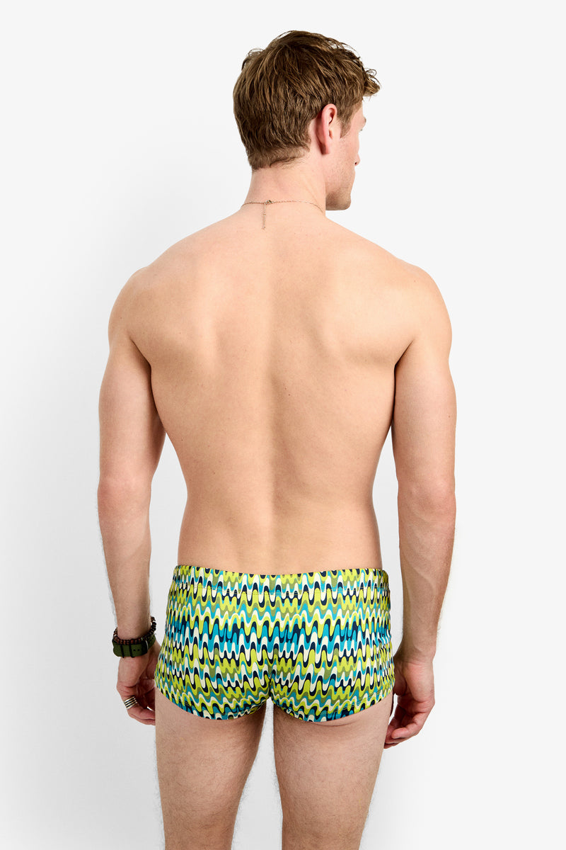 A man with light brown hair stands with his back to the camera, wearing the Sorrento Swim Trunk – Psychedelic Voyage, featuring a bold yellow, green, and blue zigzag pattern. The background is plain white.