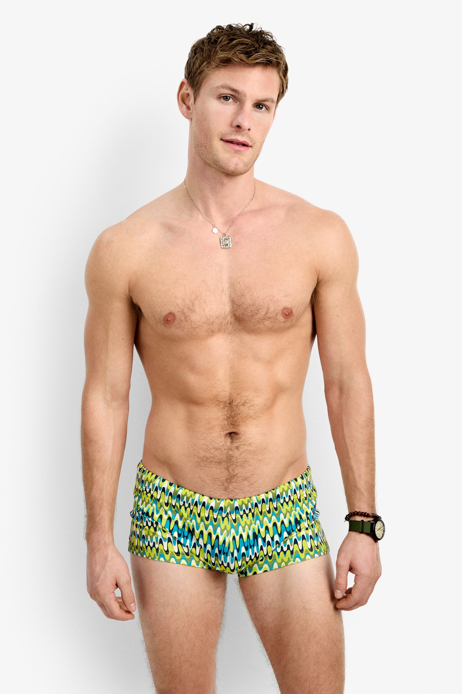 A shirtless man models the Sorrento Swim Trunk - Psychedelic Voyage against a plain white background, accessorized with a wristwatch, bracelet, and necklace while looking at the camera.