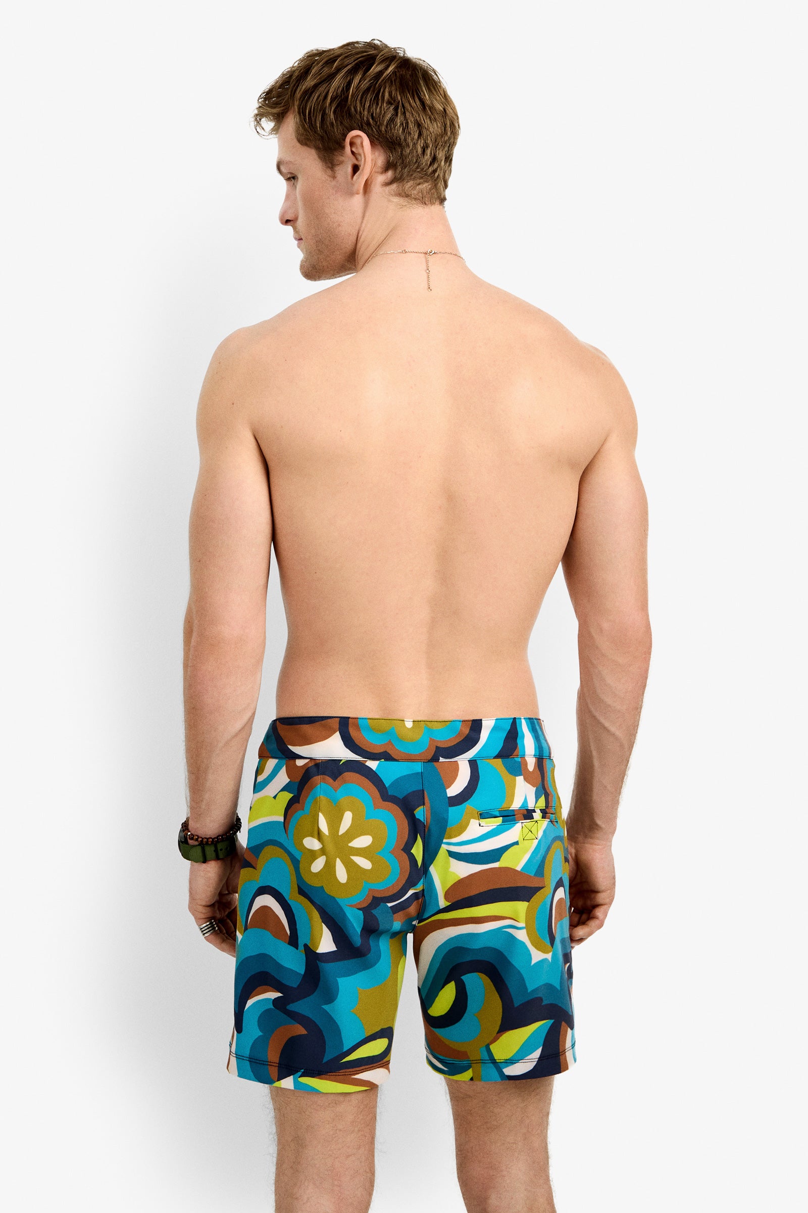 A shirtless man stands with his back to the camera, wearing Honolulu Board Short - Fonda Floral swim trunks with a bold, abstract floral pattern in blue, green, yellow, and brown—ideal for a summer vacation.