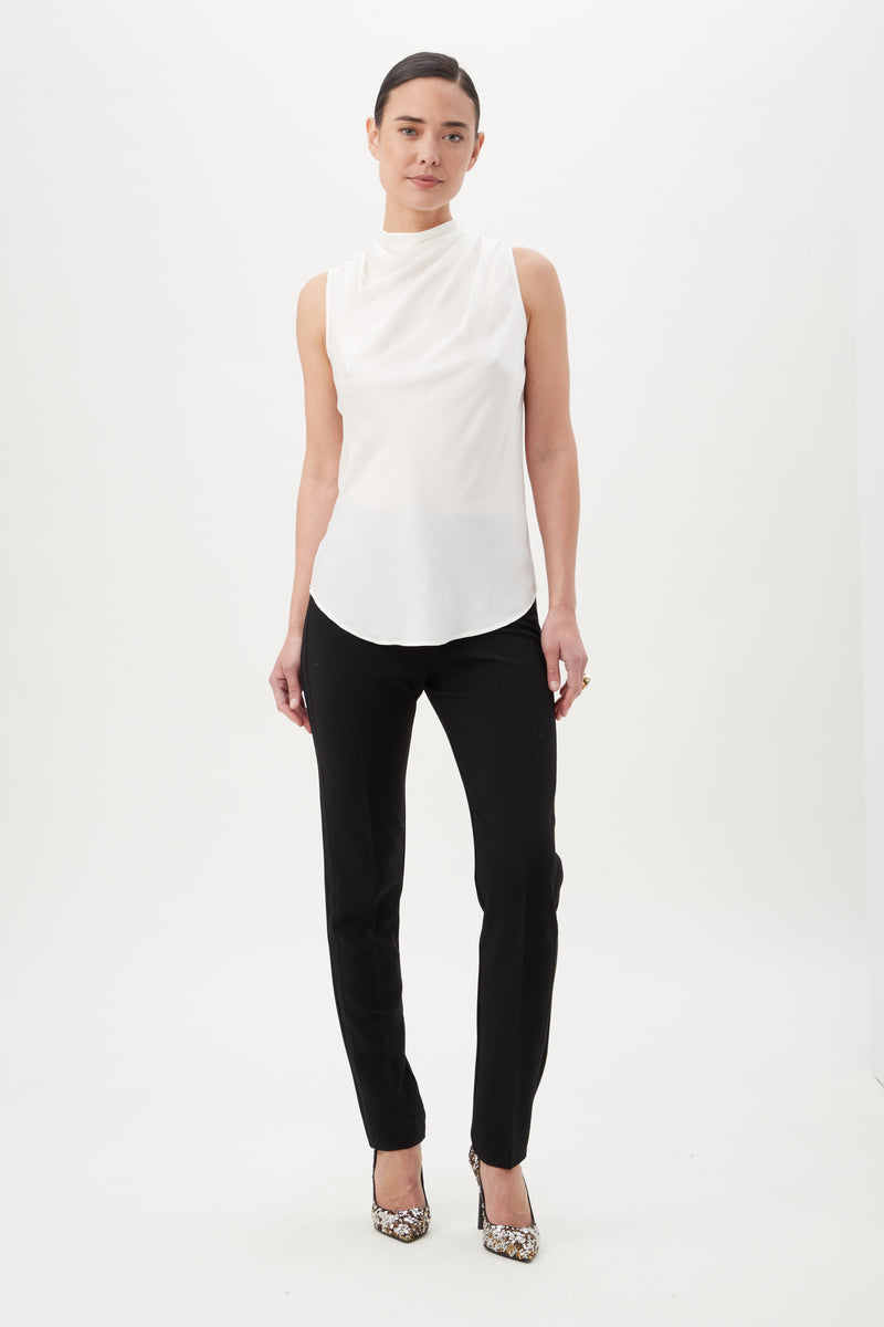 A woman stands against a white background wearing the sleeveless Lenaya Top with a funnel neckline, black pants, and patterned high heels. Her hair is pulled back and her arms rest relaxed at her sides.